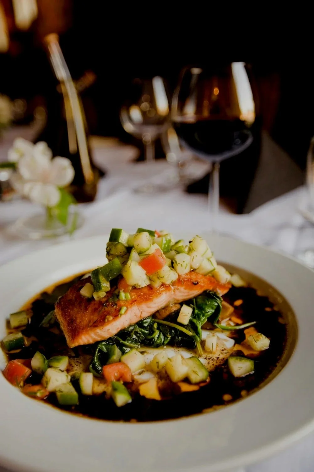 A plate with cooked salmon topped with diced cucumber and tomato, served with a dark sauce, and accompanied by wilted greens, on a formal dining table with glasses of red wine in the background.
