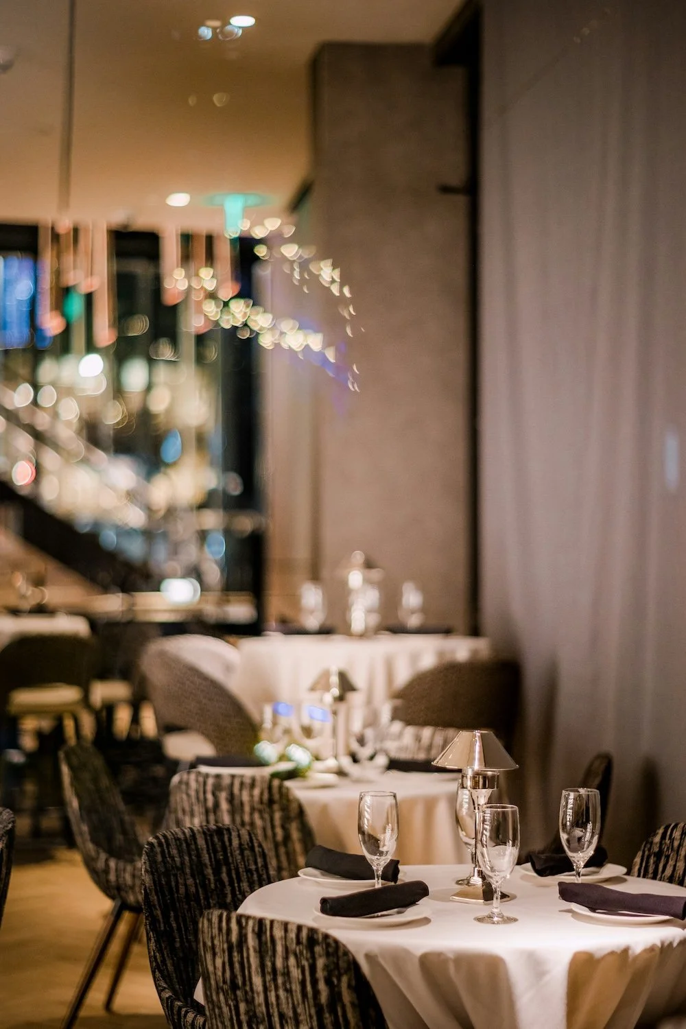 Elegant restaurant dining area with round tables, black napkins, wine glasses, and patterned chairs, softly lit with small table lamps and blurred city lights in the background.