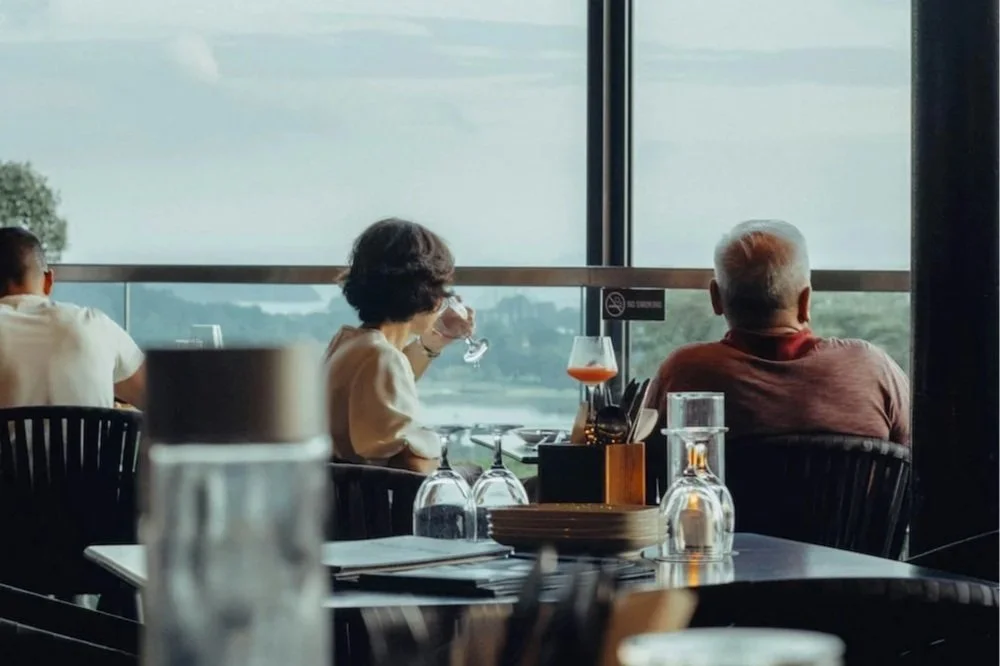 People dining at a restaurant with large windows showing a river and trees outside