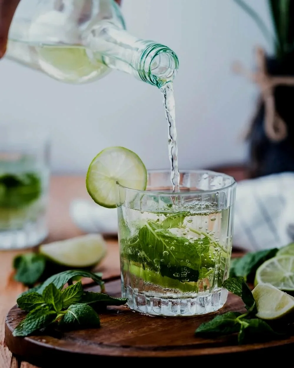 A glass of mojito being filled with sparkling water, garnished with a lime slice, fresh mint leaves, and lime wedges on a wooden board.