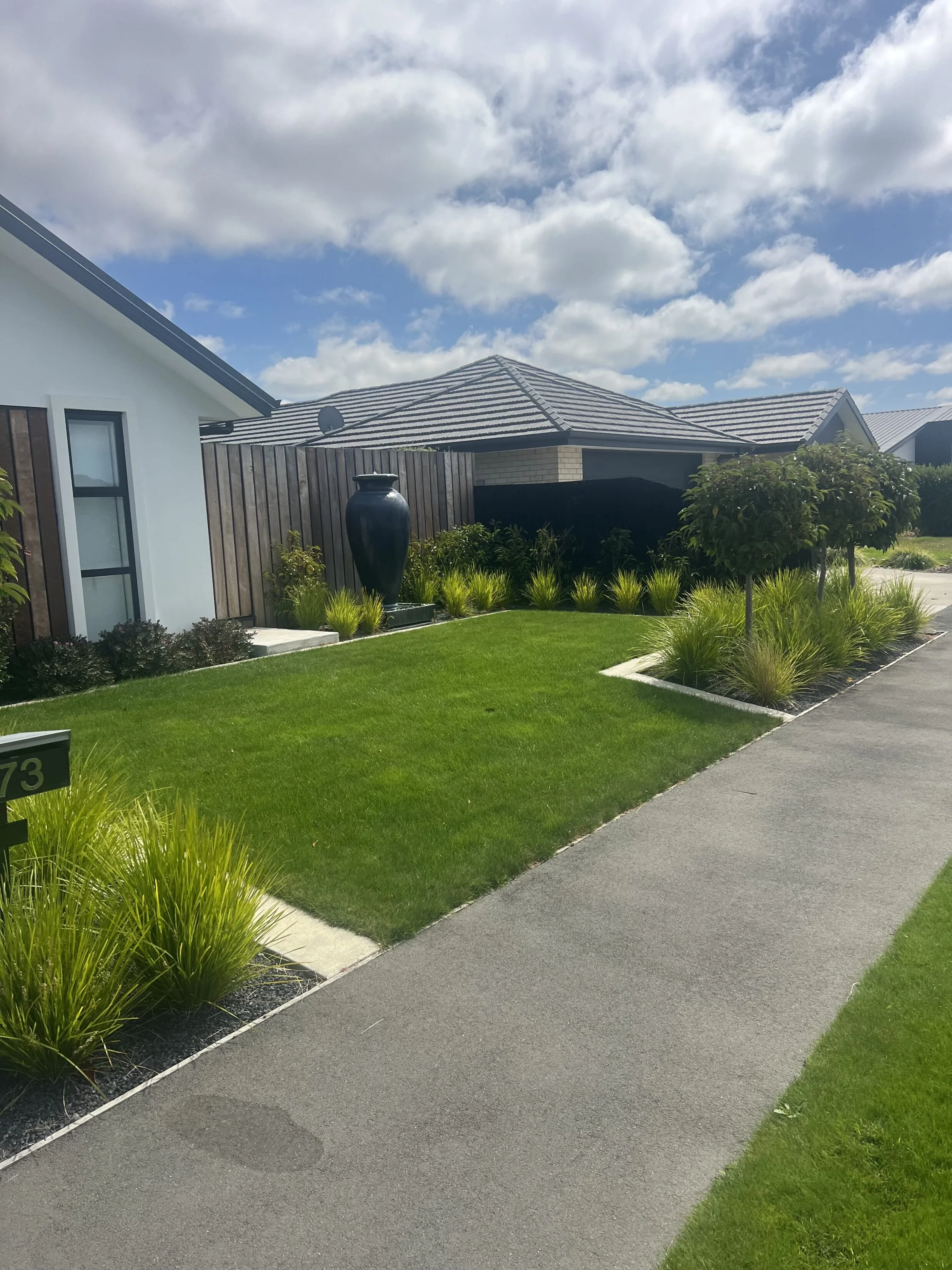 Front yard with green grass, landscaped plants, and concrete sidewalk in a suburban neighborhood under a partly cloudy sky.