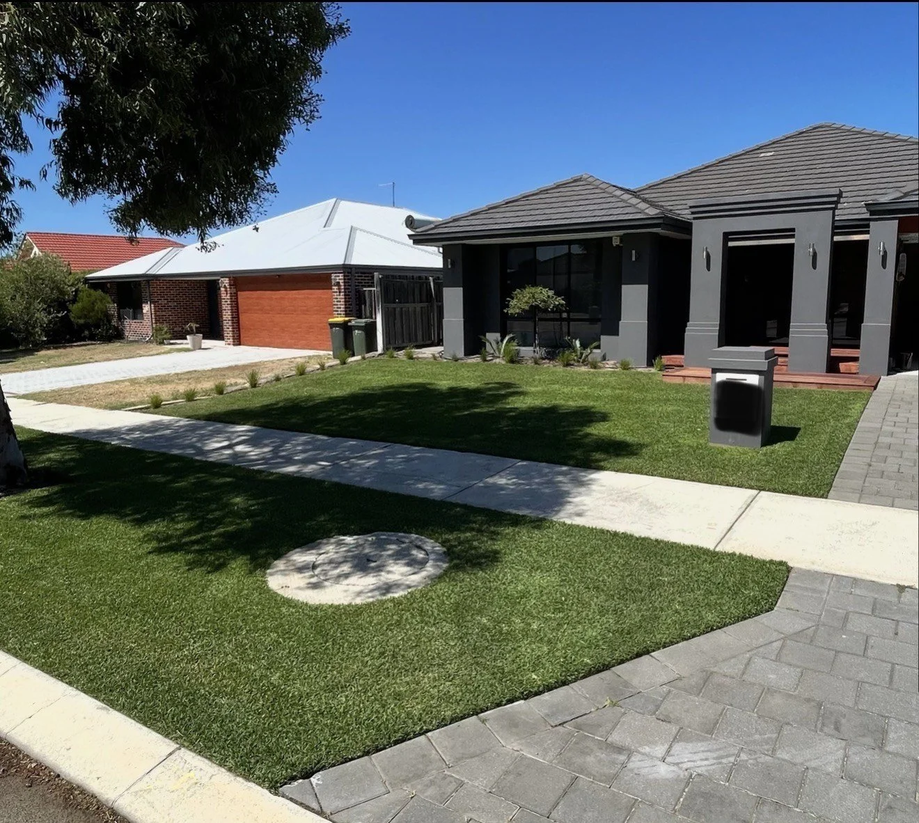 Front yard of a modern house with green lawn, concrete walkway, and a gray and black exterior. The house has a driveway, a mailbox, and some small plants near the windows. A tree is casting a shadow on the lawn.