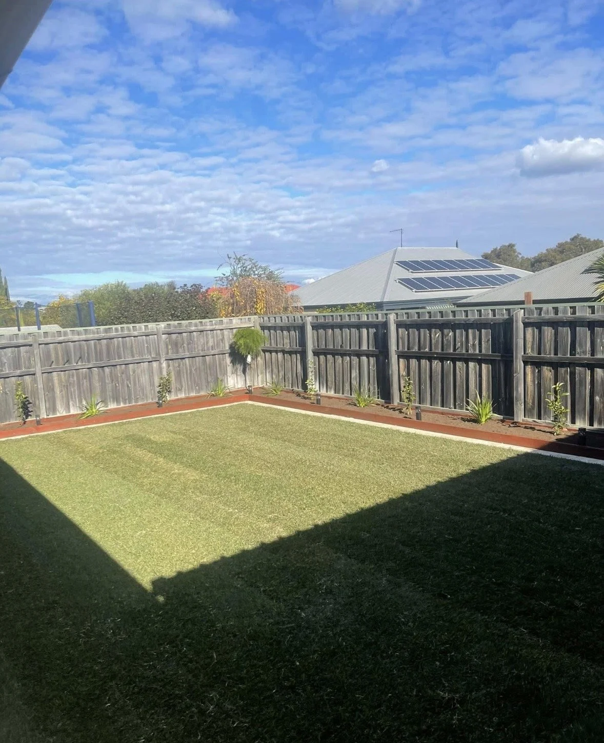 A backyard with a well-maintained lawn, surrounded by a wooden fence, with a house in the background that has solar panels on the roof, and trees with blue skies and scattered clouds overhead.