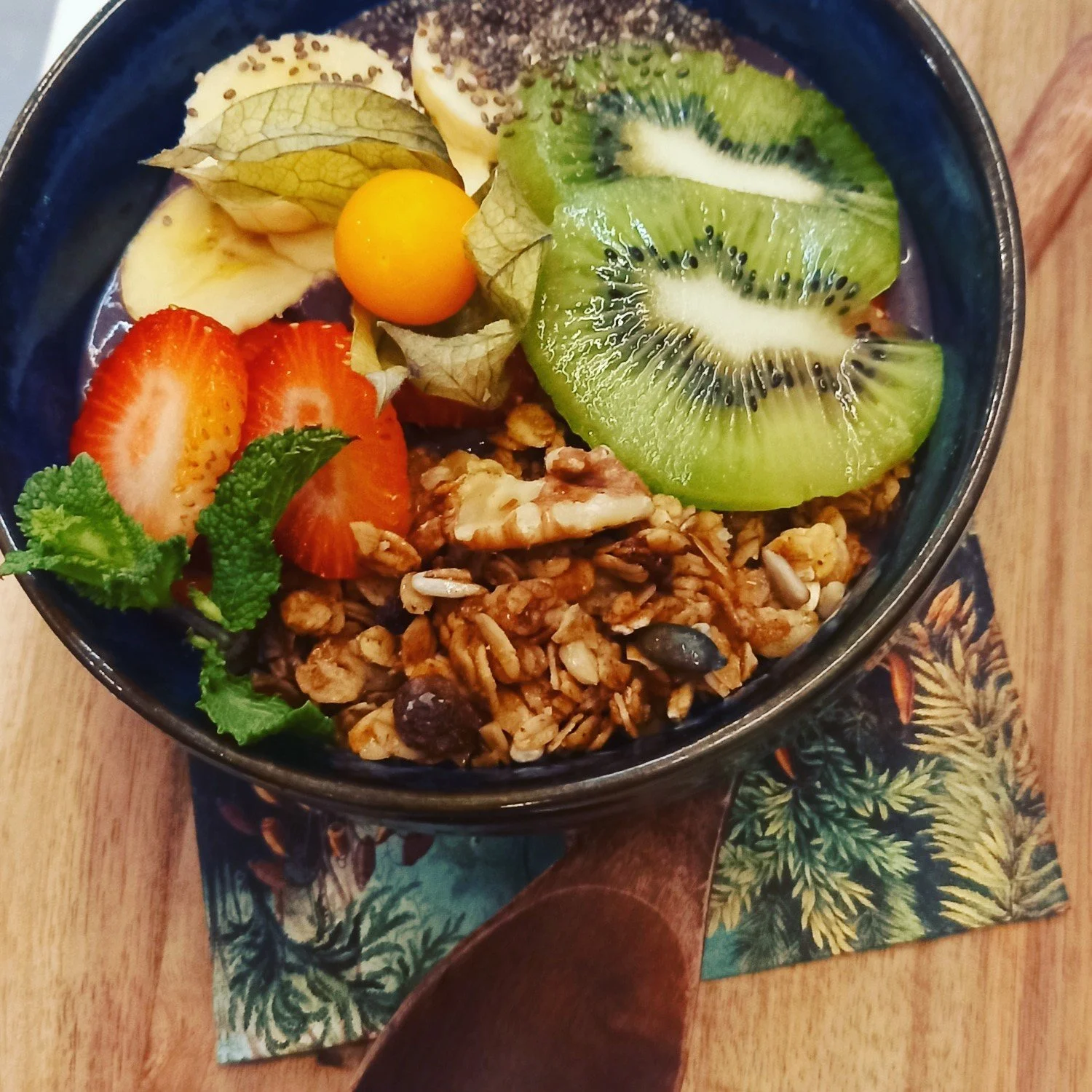 A bowl of granola with sliced strawberries, kiwi, and banana, topped with a physalis and a sprig of mint, on a wooden table with a decorative napkin underneath.