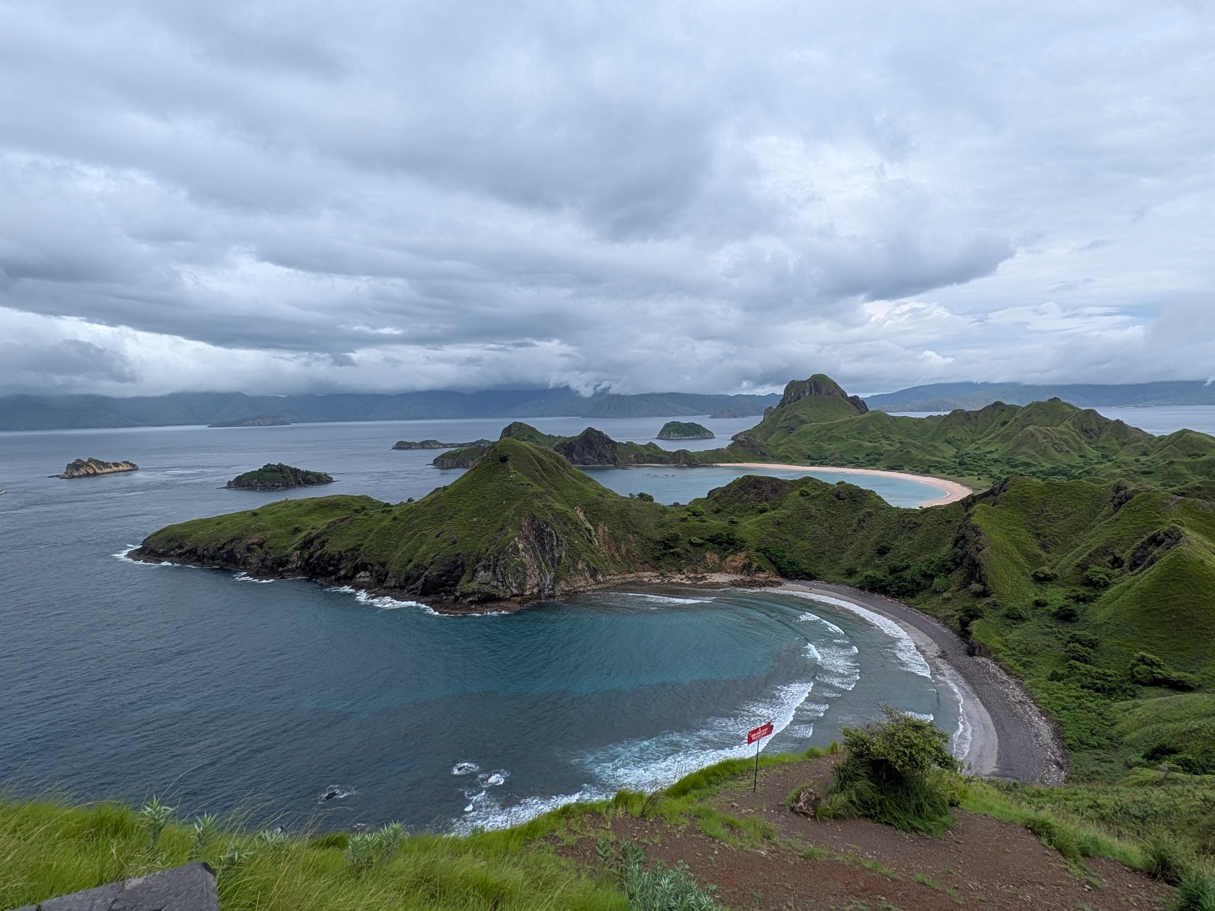 A coastal landscape with green hills and islands in the distance, a curved beach with dark sand, and a cloudy sky overhead.
