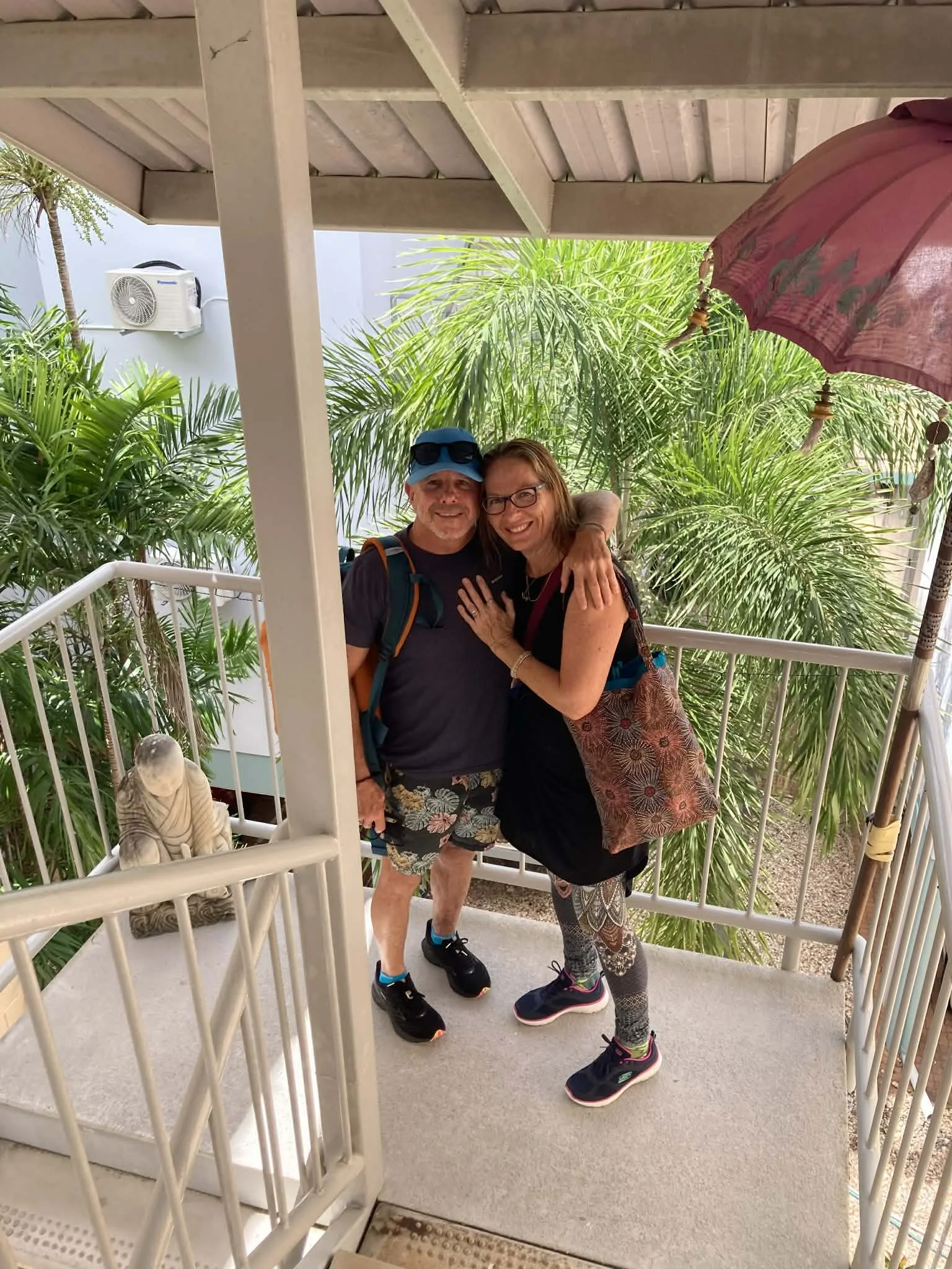 A smiling man and woman hugging on a balcony surrounded by lush tropical plants, a pink parasol hanging above them.