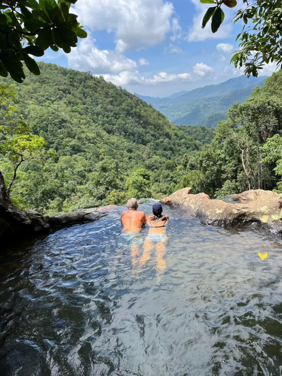 Two people, a man and a woman, sitting in a natural pool overlooking a lush green mountain landscape on a sunny day.