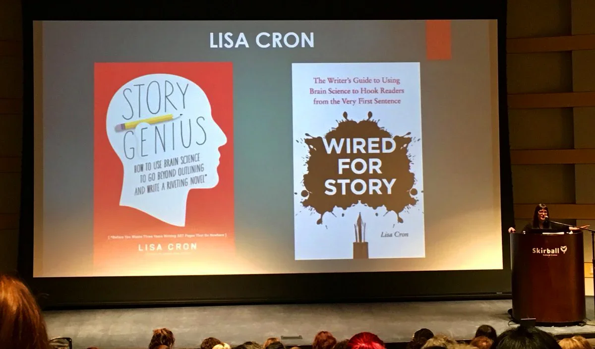 Presentation slide displaying two book covers by Lisa Cron, titled "Story Genius" and "Wired for Story," with a speaker standing at a podium in an auditorium filled with audience members.