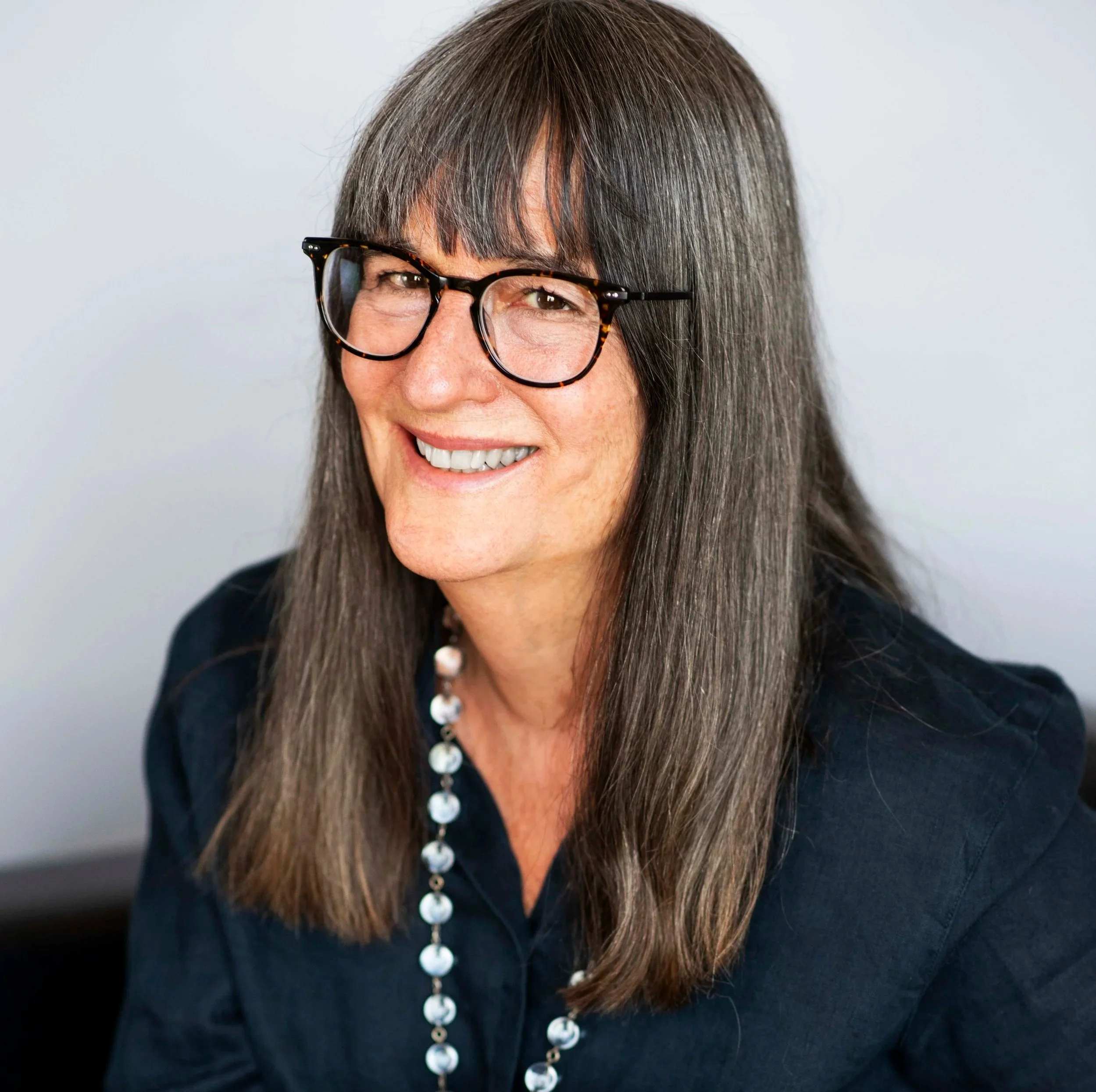 Close-up of a smiling middle-aged woman with long gray hair, black glasses with tortoiseshell pattern, silver beaded necklace, and black shirt against a plain background.