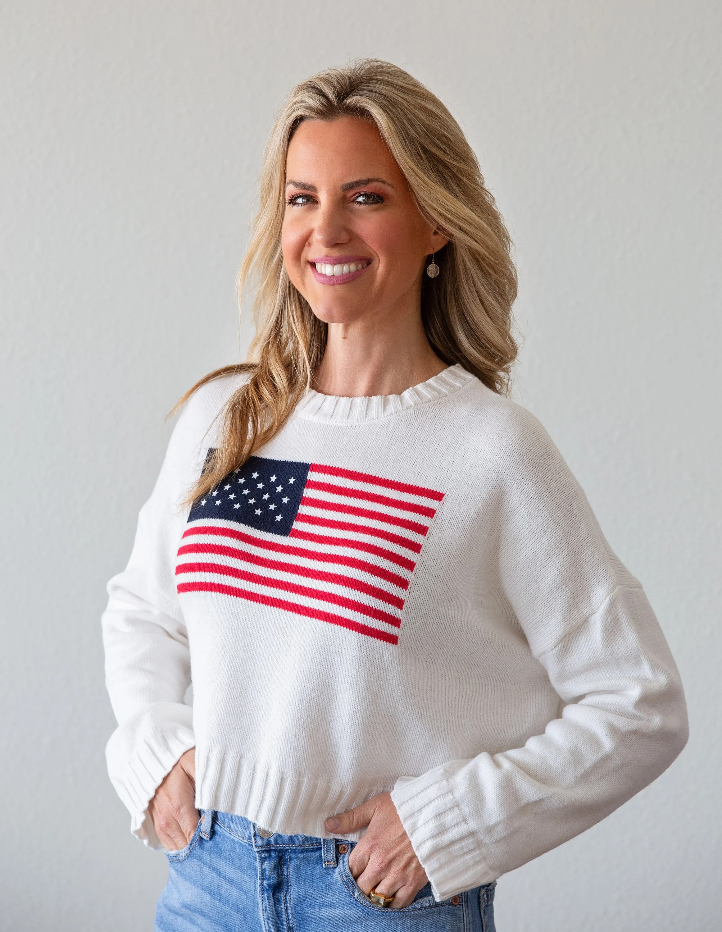 A smiling woman with blonde hair wearing a white sweater with an American flag graphic and blue jeans, standing against a plain light gray background.
