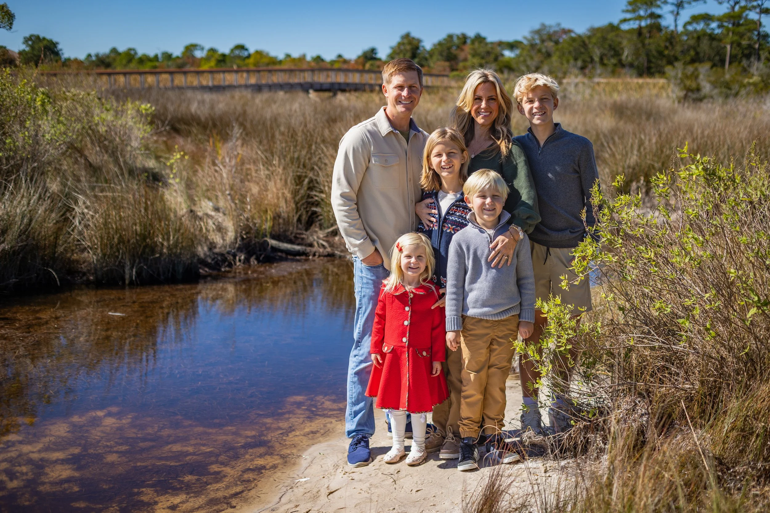 A family of six standing on a sandy trail near a creek in a natural park setting, with trees and a wooden bridge in the background on a sunny day.