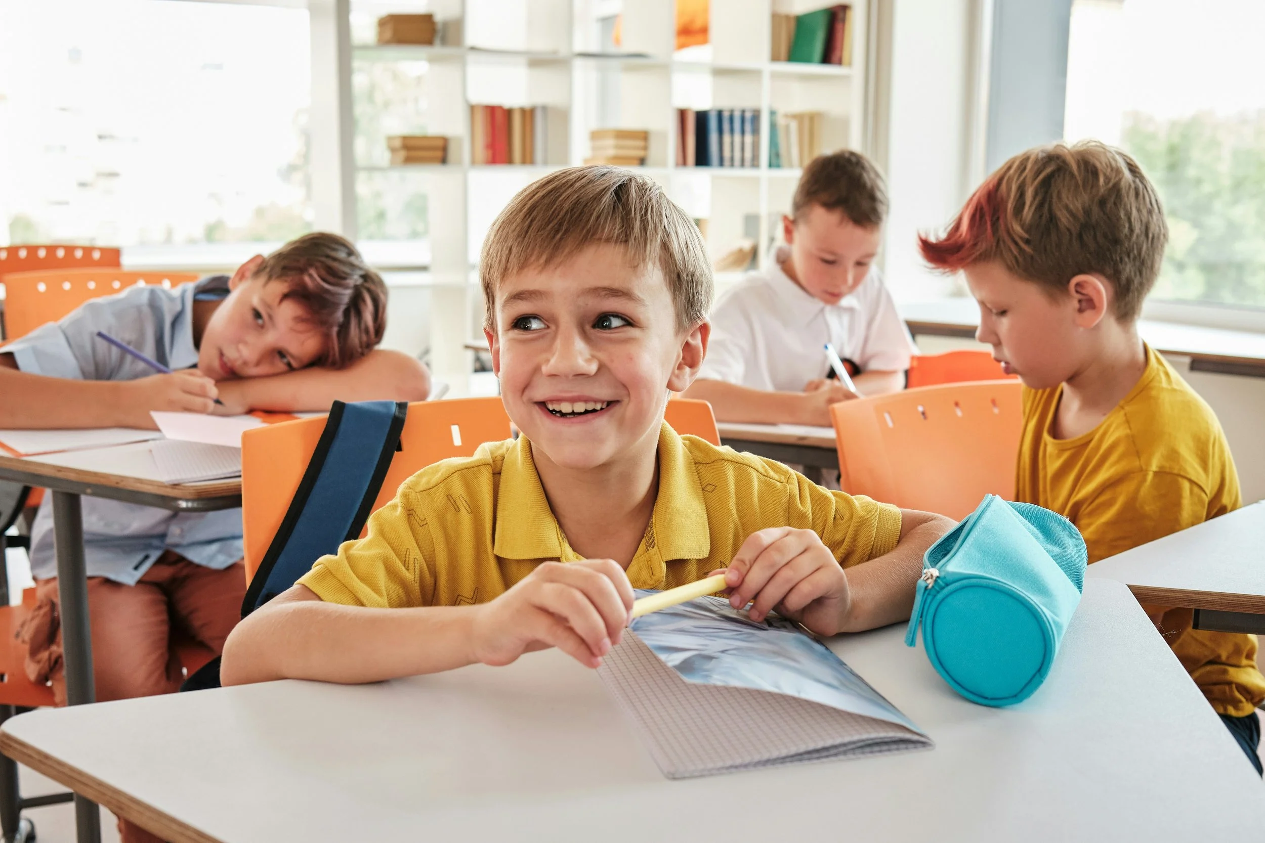 Young boys sitting at desks in a classroom, some writing, one smiling and looking to the side.