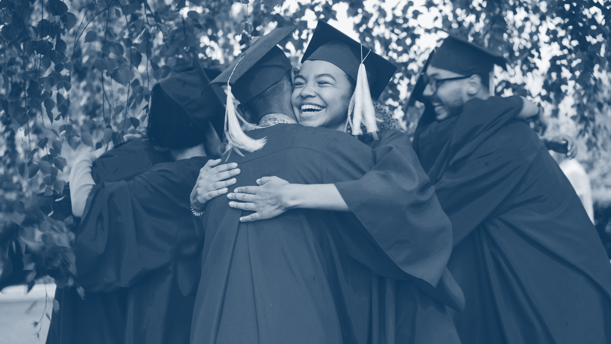 Group of students in caps and gowns hugging, celebrating graduation outdoors with trees in the background.