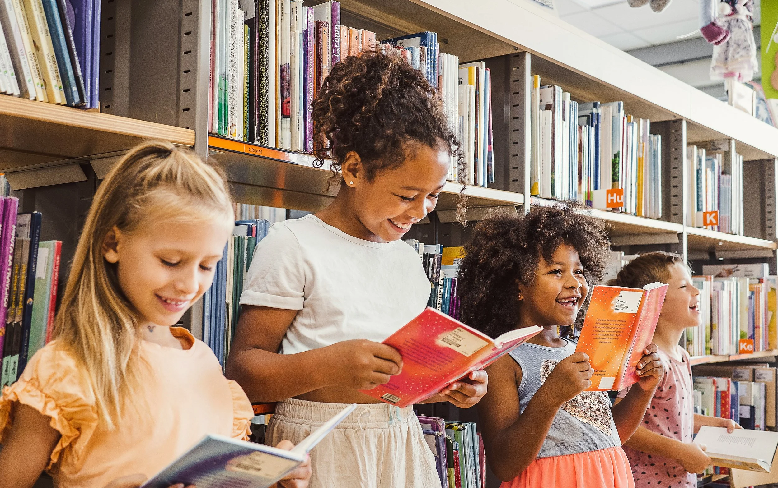 A group of children reading books in a library, standing in front of bookshelves filled with colorful books.