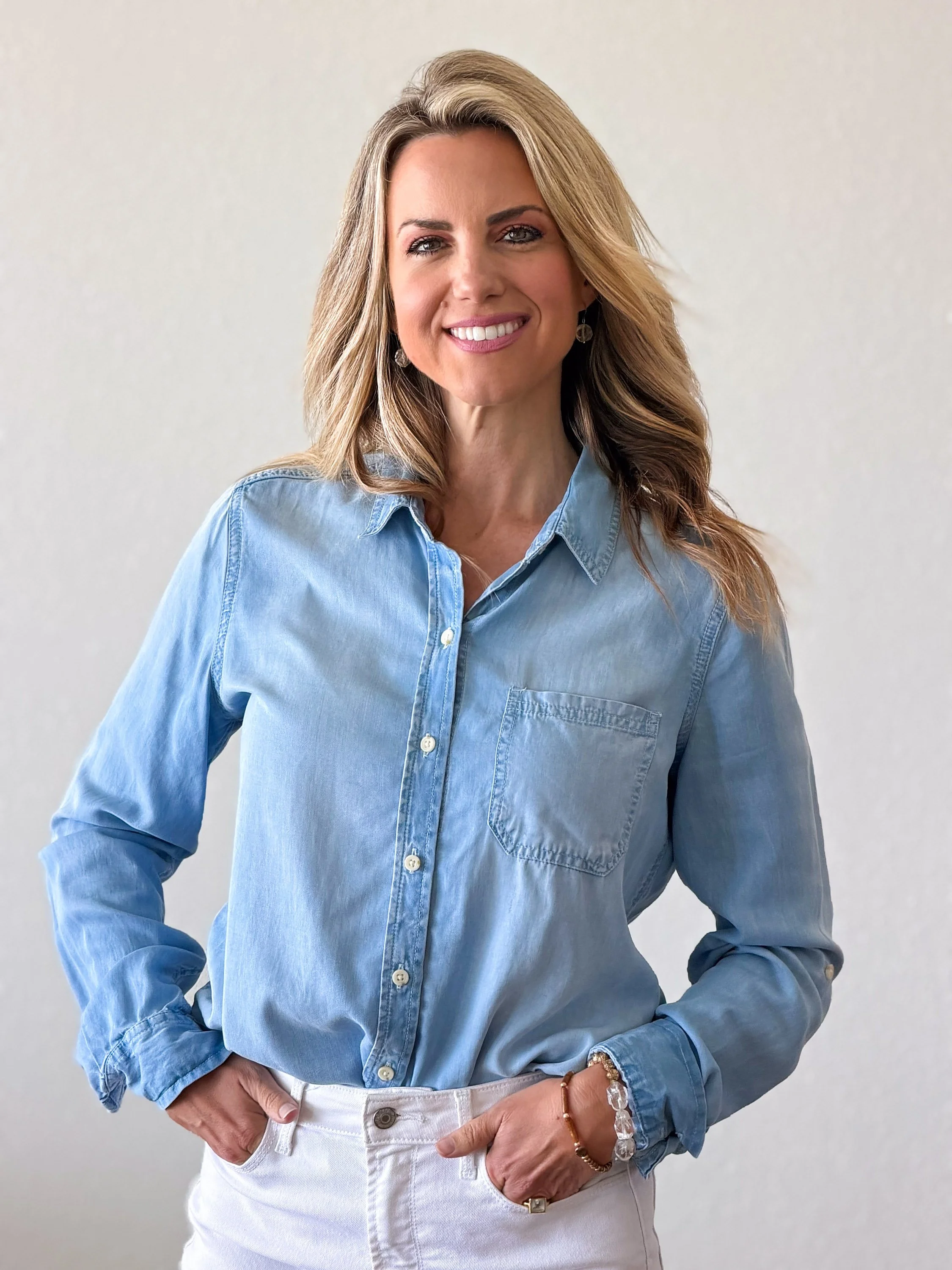 A woman with long blonde hair wearing a light blue denim shirt with rolled up sleeves, white jeans, and jewelry, standing against a plain white background.