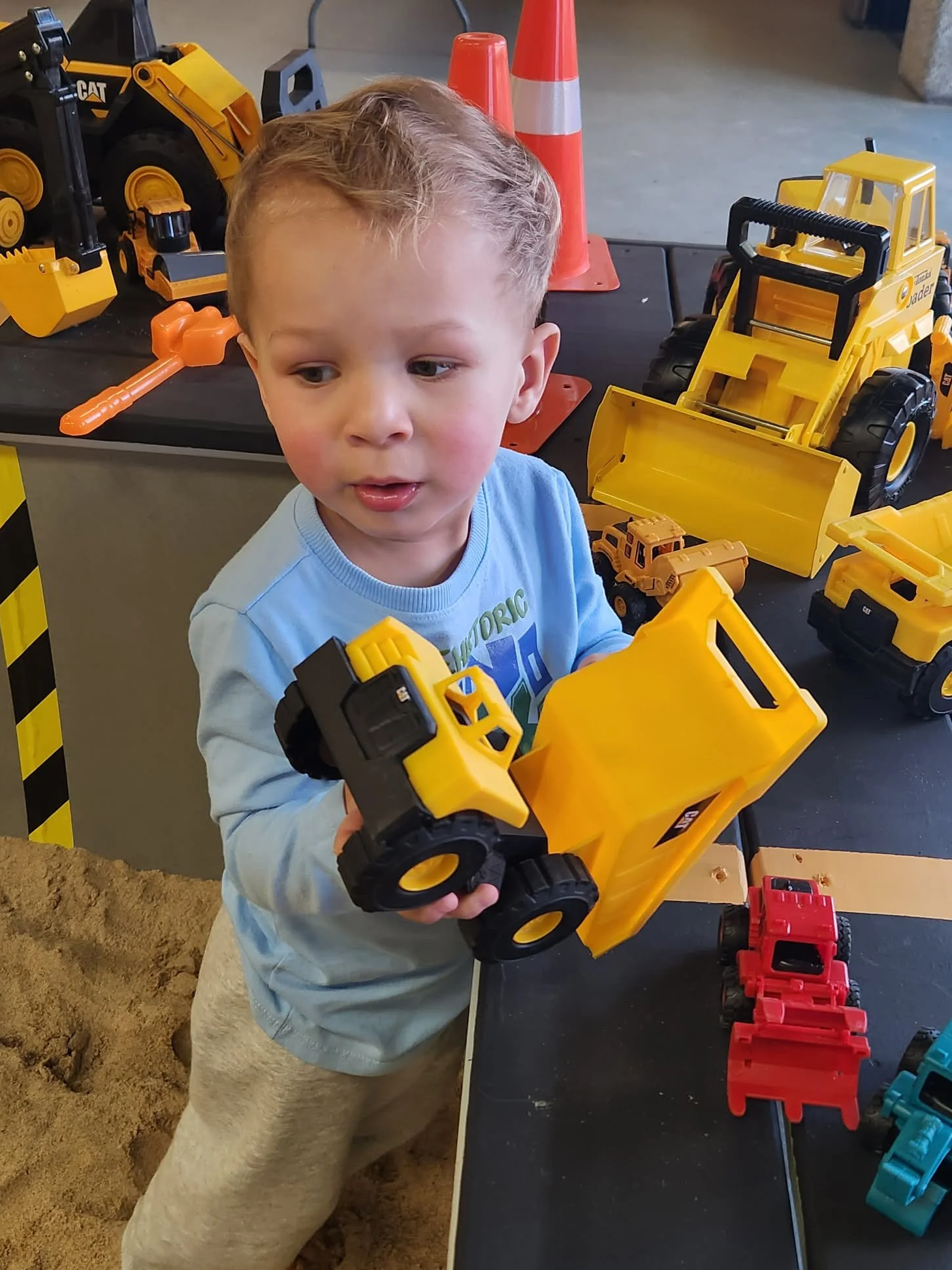 A young boy with blonde hair wearing a light blue shirt and beige pants playing with yellow construction toy trucks at a sandbox.