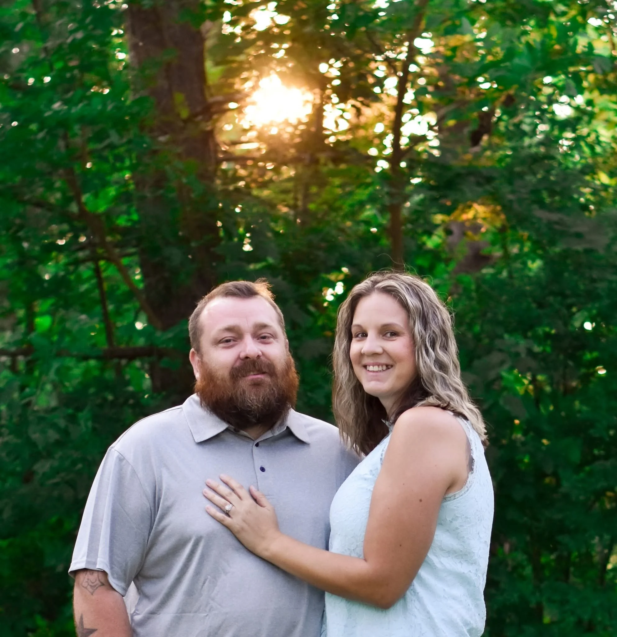 A couple standing outdoors in front of trees with sunlight shining through, the woman is wearing a white sleeveless dress and has her hand on the man's chest, both are smiling.