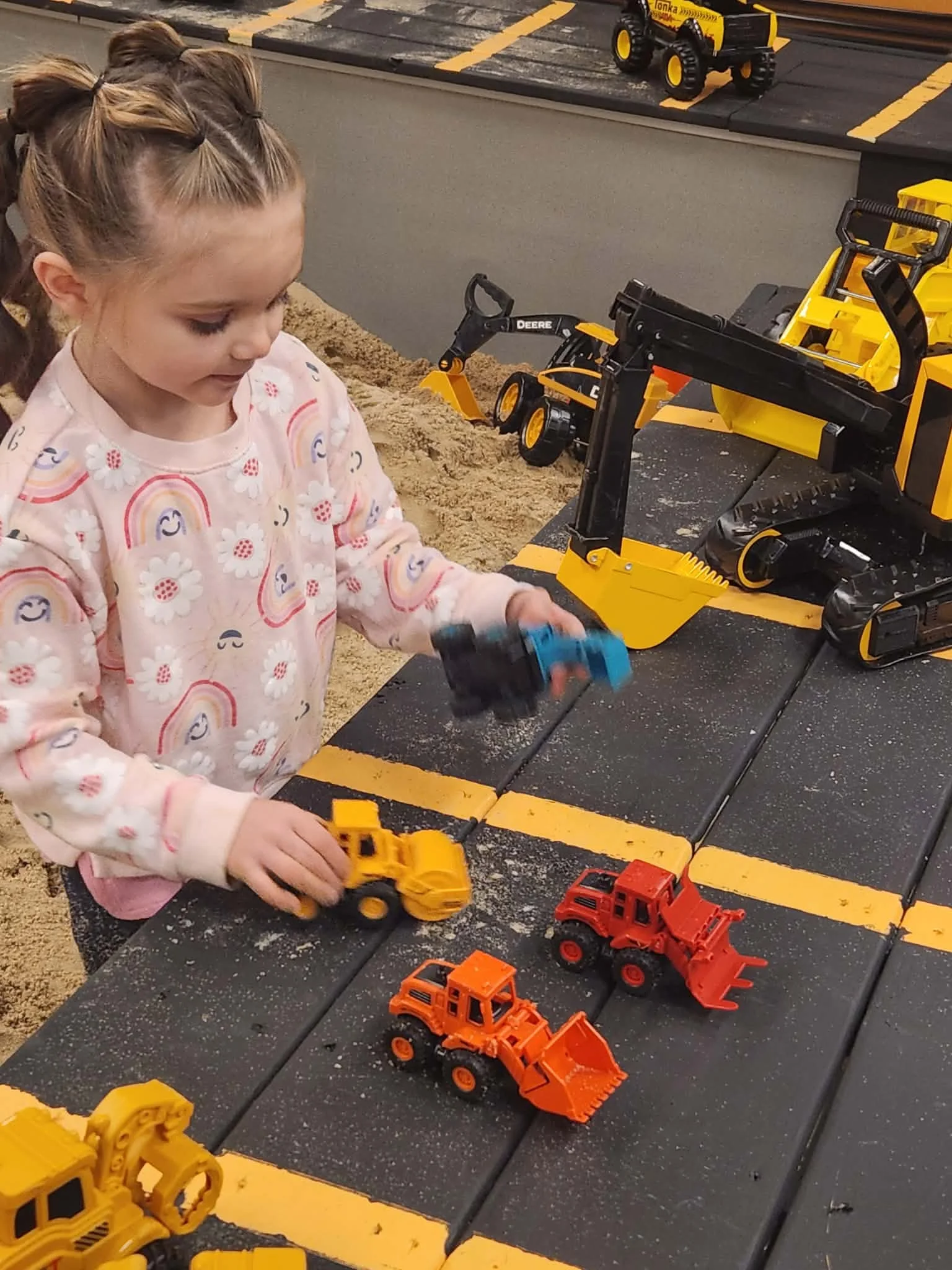 Young girl playing with toy construction vehicles in a sandbox with a construction-themed play area.