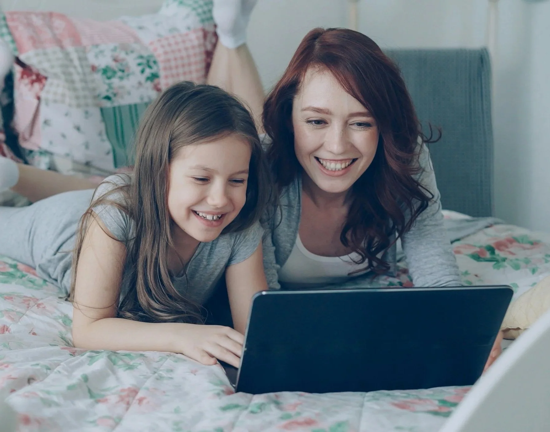 A smiling woman and a girl lying on a bed looking at a laptop screen.