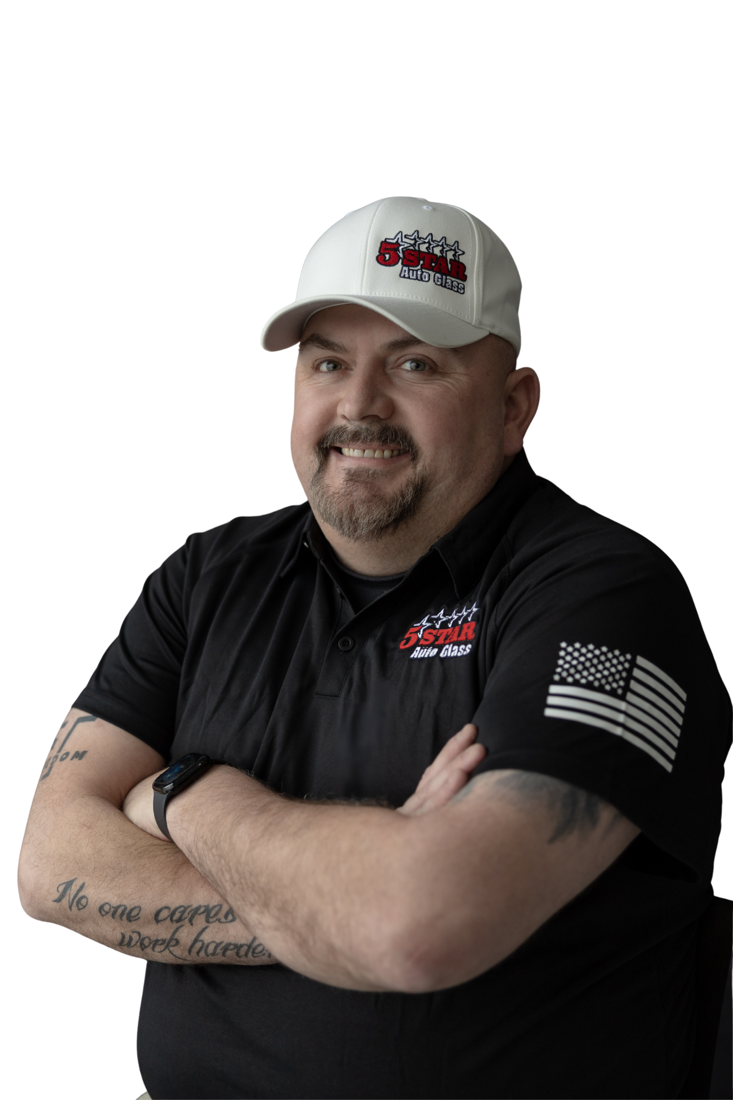A man with a beard and mustache, wearing a white baseball cap and black polo shirt with a logo, standing with arms crossed, smiling against a plain black background.