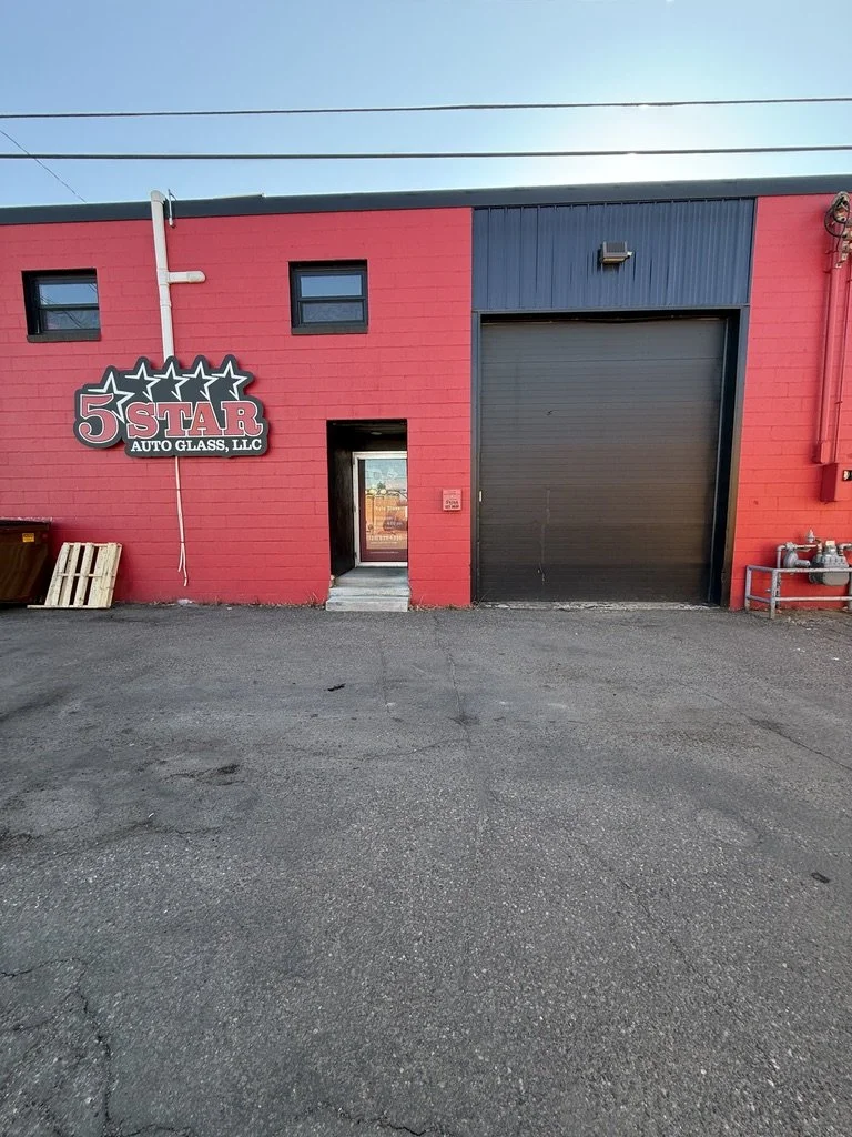 Red building with a sign that reads '5 Star Auto Glass, LLC' on the left, a small door in the center, and a large black garage door on the right. The ground in front is paved asphalt.