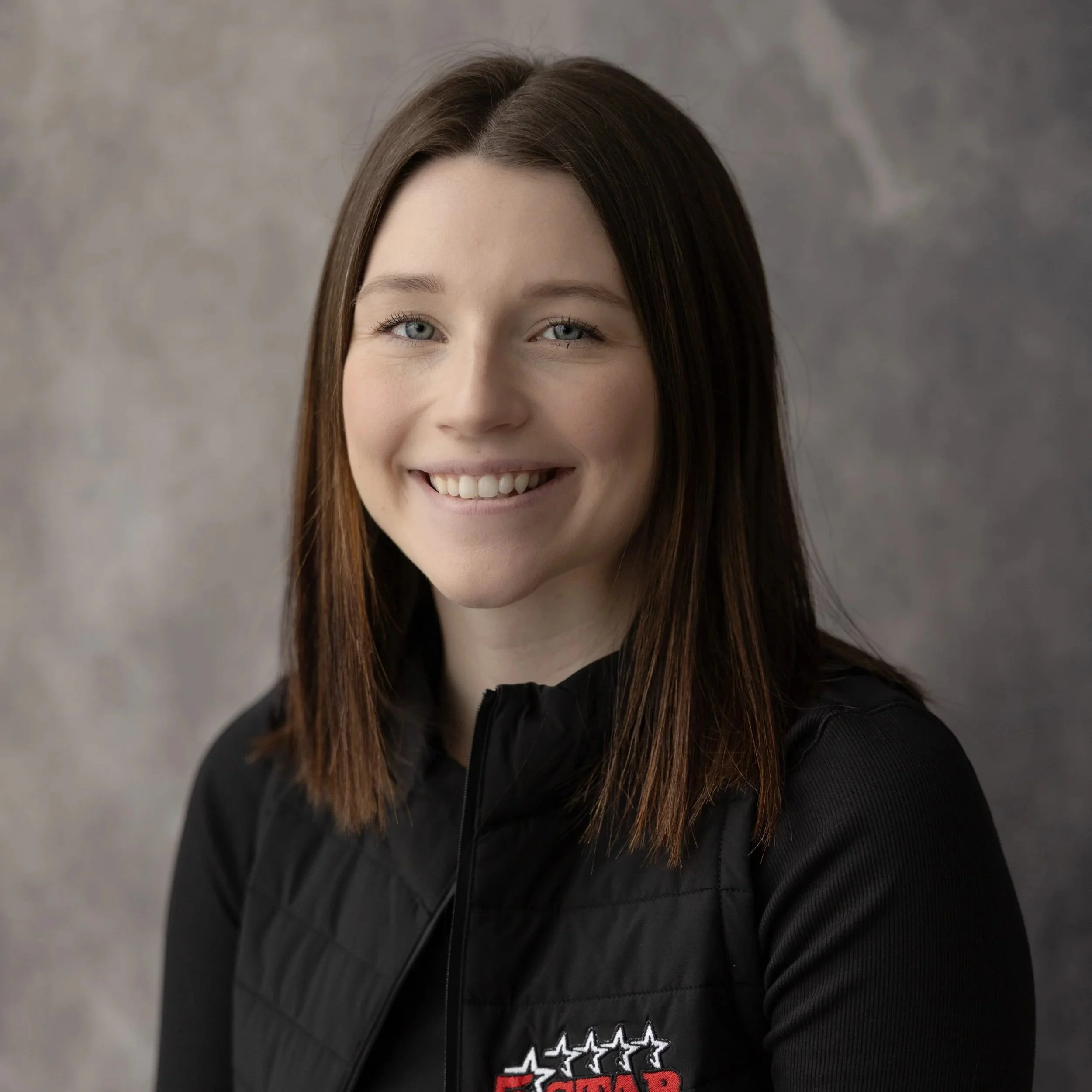 Headshot of a young woman with shoulder-length brown hair, blue eyes, and a bright smile, wearing a black jacket with a logo.