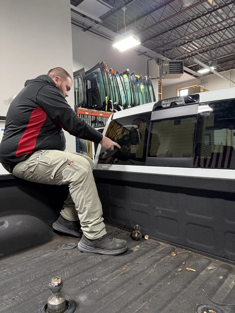 A man sitting in the bed of a pickup truck inside a warehouse, touching the truck's rear window, with glass panels stacked on a shelf in the background.