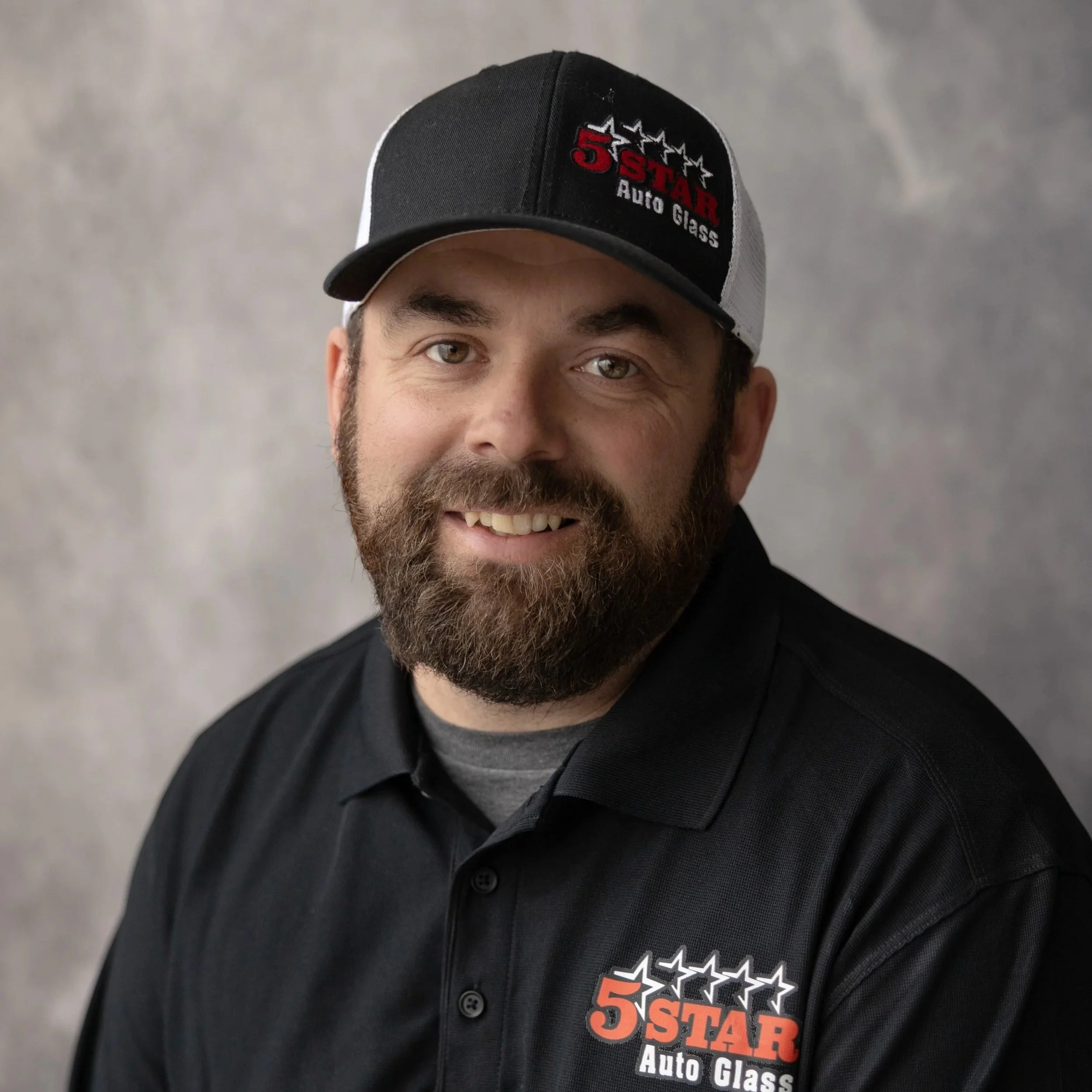A man with a beard and mustache wearing a black polo shirt with a logo that reads '5 Star Auto Glass' and a black and gray cap with the same logo, smiling against a gray background.