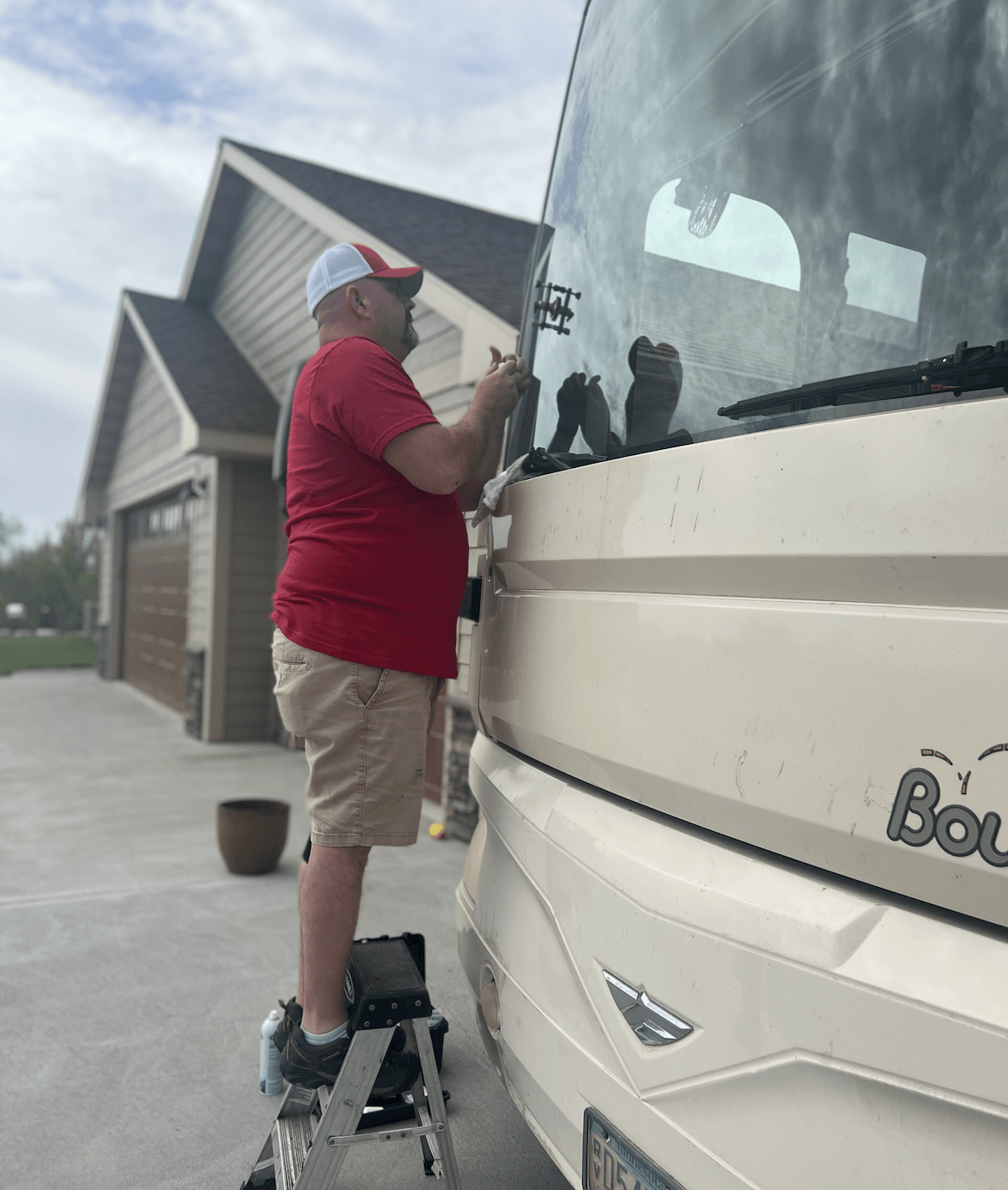 A man in a red shirt, khaki shorts, and a white cap is standing on a step stool cleaning or repairing the front of a white boat parked in a driveway. The background shows a house with a garage and a cloudy sky.