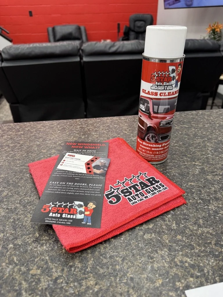 A can of glass cleaner, a red cloth with logo, and a flyer on a speckled countertop in a waiting area with black chairs and red brick wall.