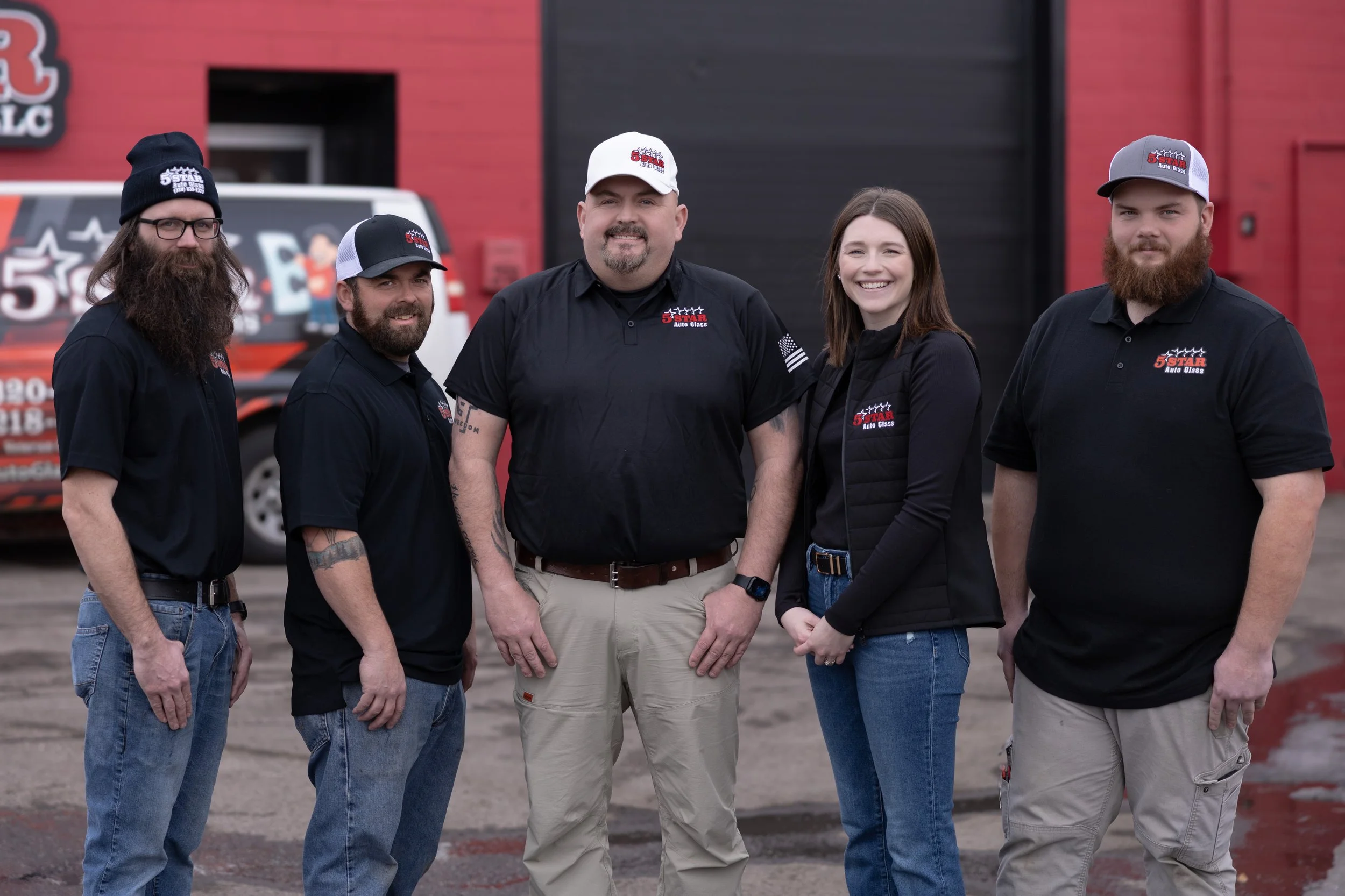 Family or team of five people standing in front of a red and black building, posing for the photo. They are wearing black shirts with the logo '5 Star Auto Glass,' with some also wearing caps and a vest. They are smiling, and there is a van with similar branding in the background.