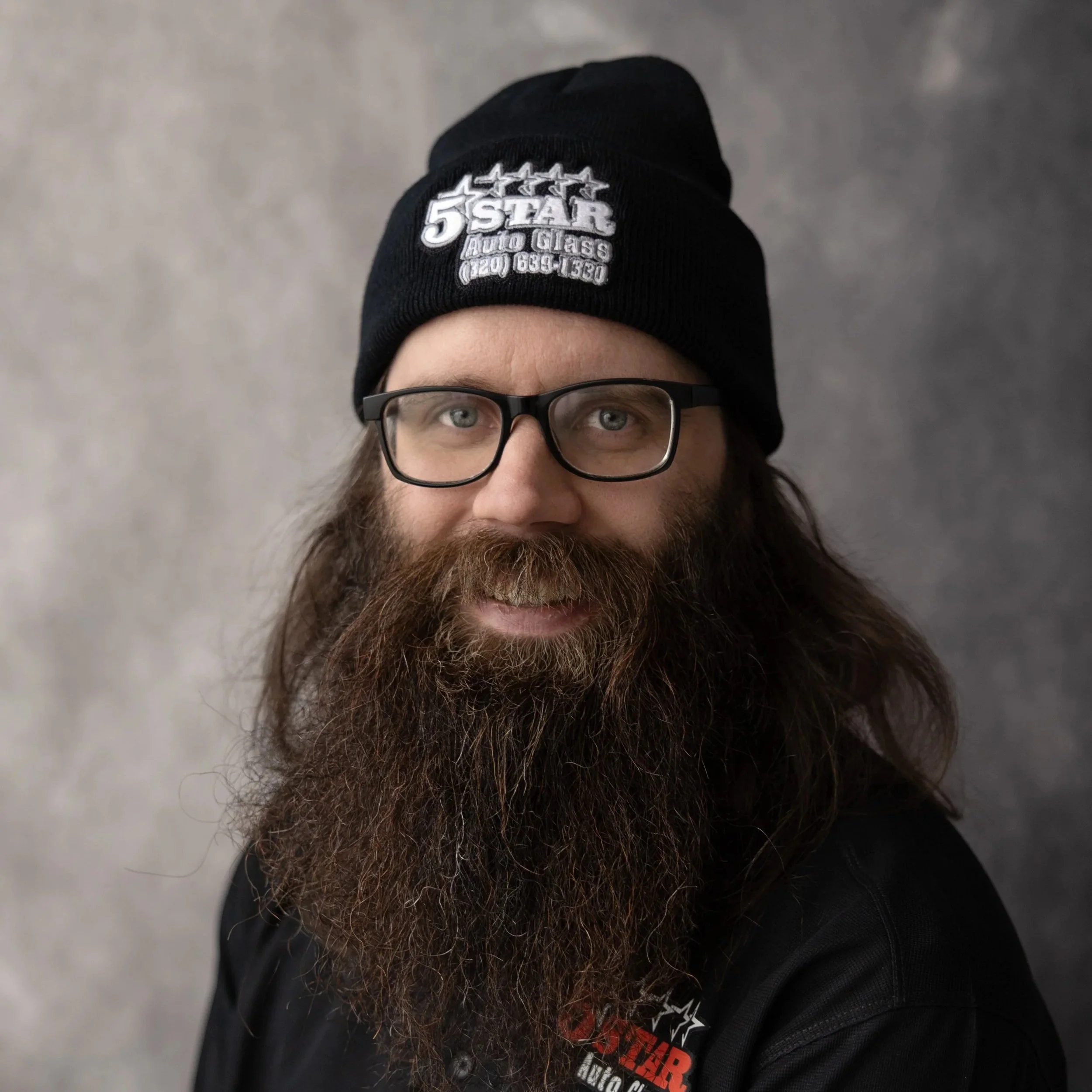 A man with long hair and a full beard wearing glasses, a black beanie with a logo, and a black shirt with a logo, smiling at the camera against a grey background.