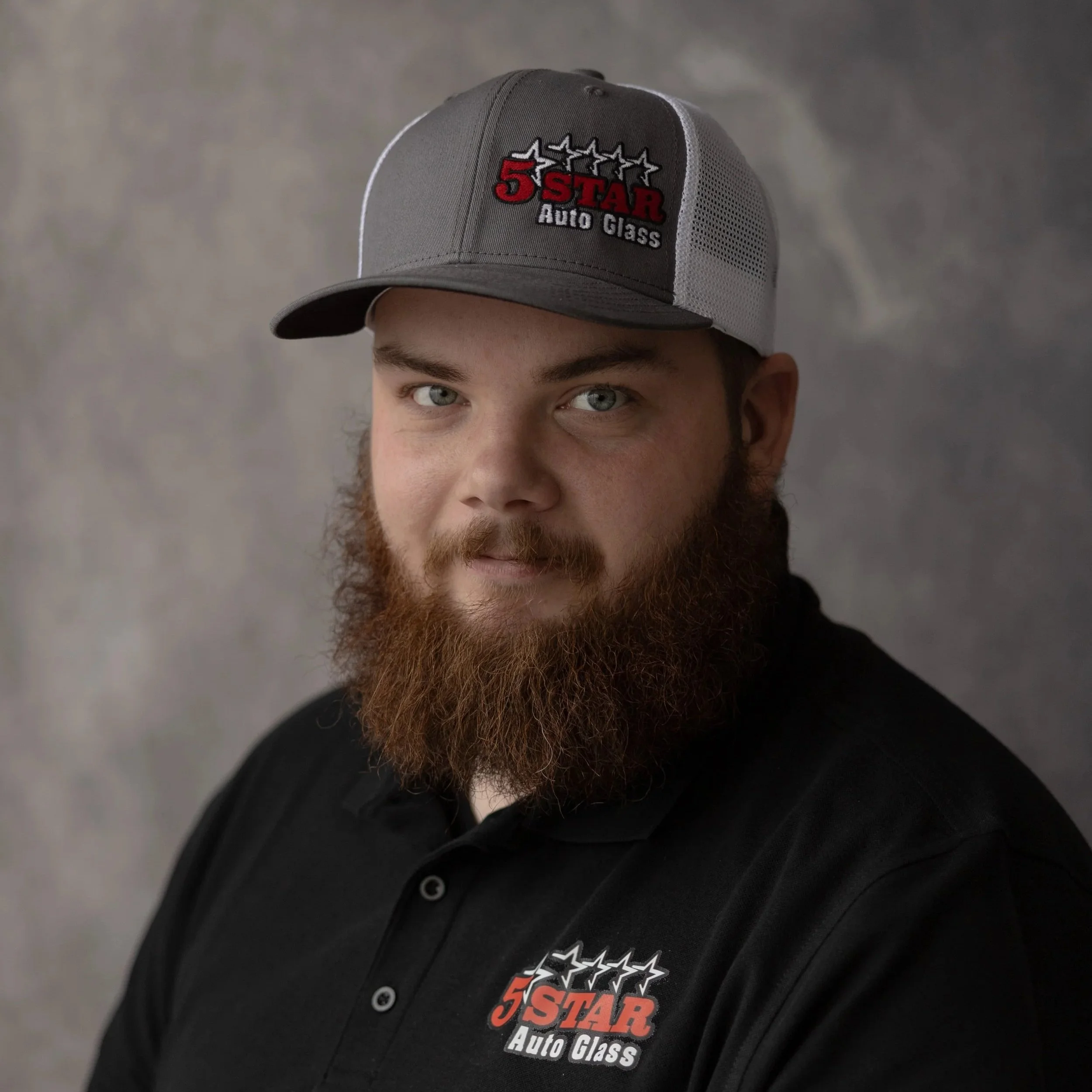 A man with a full beard wearing a gray and black baseball cap and a black polo shirt, both featuring the logo of 5 Star Auto Glass.