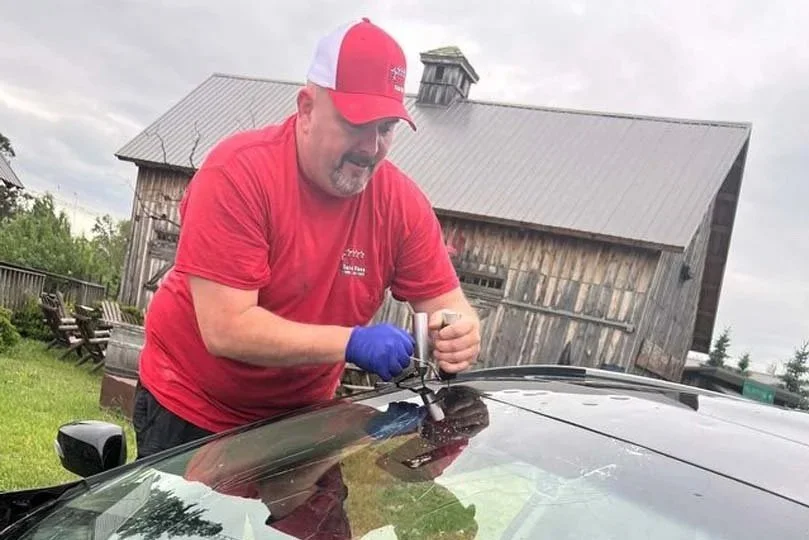 A man in a red and white cap and red shirt working on a car, using a tool while wearing blue gloves, outdoors with a rustic wooden barn in the background.