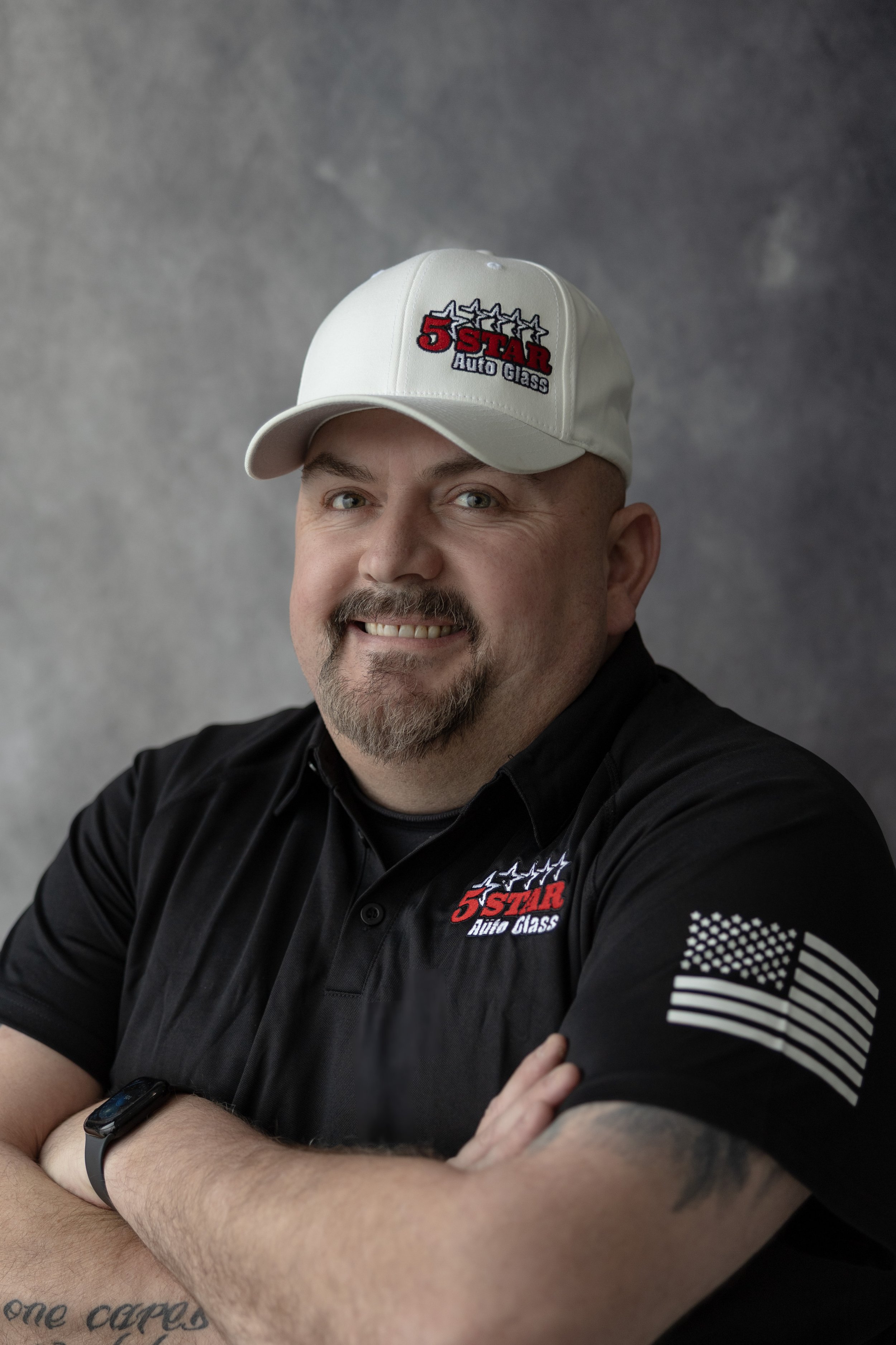 A smiling man with a goatee and short hair wearing a white cap and black polo shirt, both branded with '5 Star Auto Glass,' against a gray background.