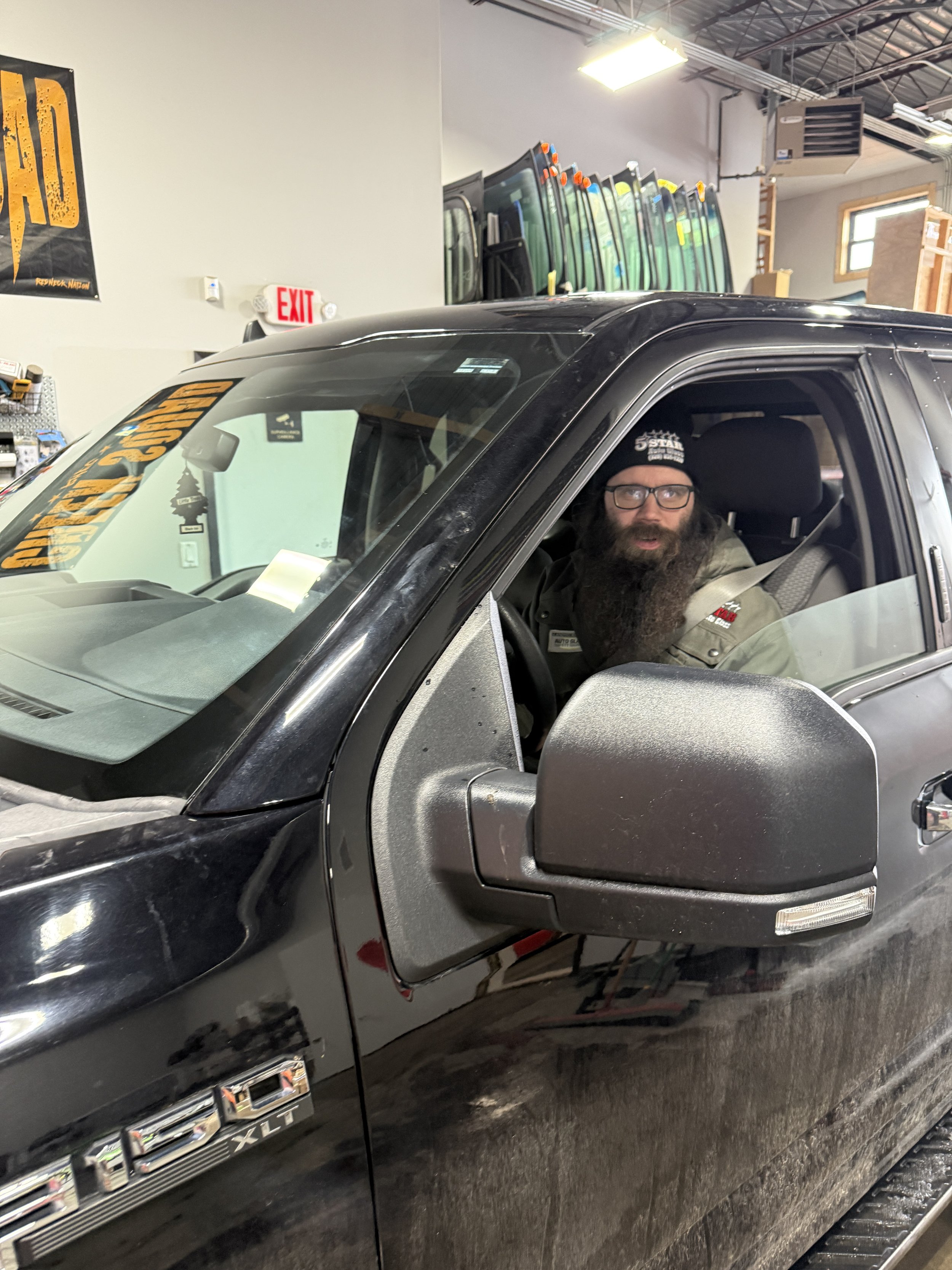 A man with a beard and glasses sitting in the driver’s seat of a black Ford F-150 truck inside a showroom or workshop, with various glass windows or doors displayed in the background.