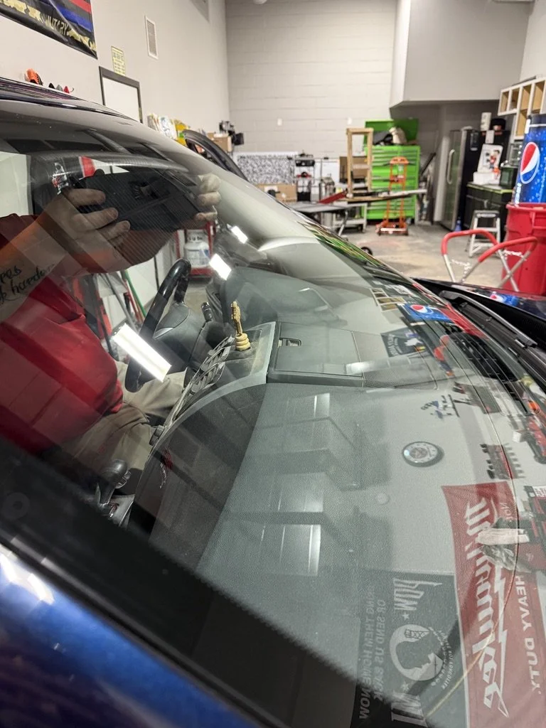 View through the windshield of a parked blue car inside a garage or workshop, with a person wearing a red or maroon shirt taking the photo. The garage contains tools, a green tool chest, a workbench, and various equipment.