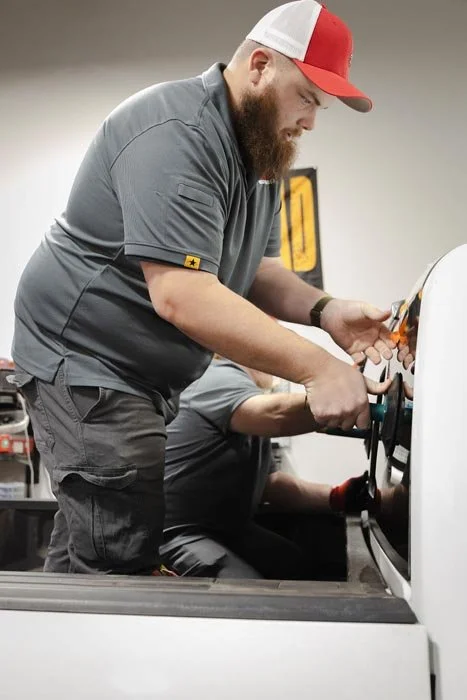 A man with a beard wearing a gray shirt and a red and white cap is working on a car, using tools at a workshop or garage.