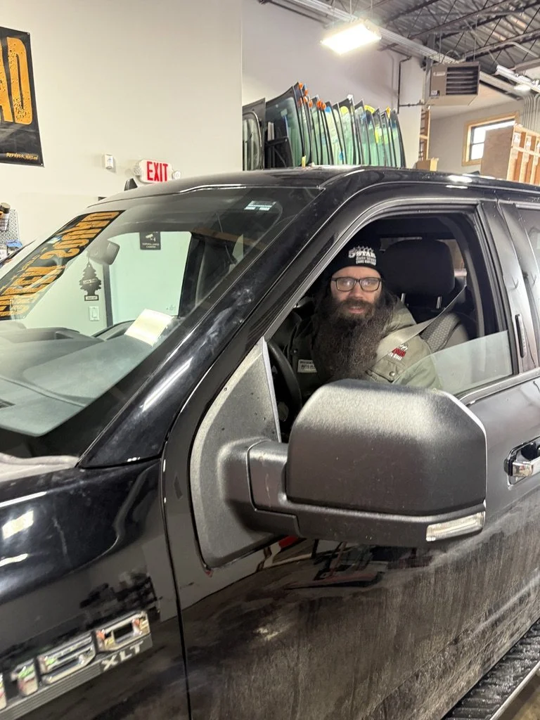 A bearded man with glasses and a black beanie sitting in the driver's seat of a black pickup truck in a warehouse or parts store, with auto glass panels stacked in the background.