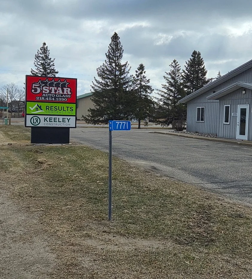 A blue street sign with the number 7771 in front of a building and some trees, with a large business sign in the background advertising auto glass, results contracting, and Keeley construction.