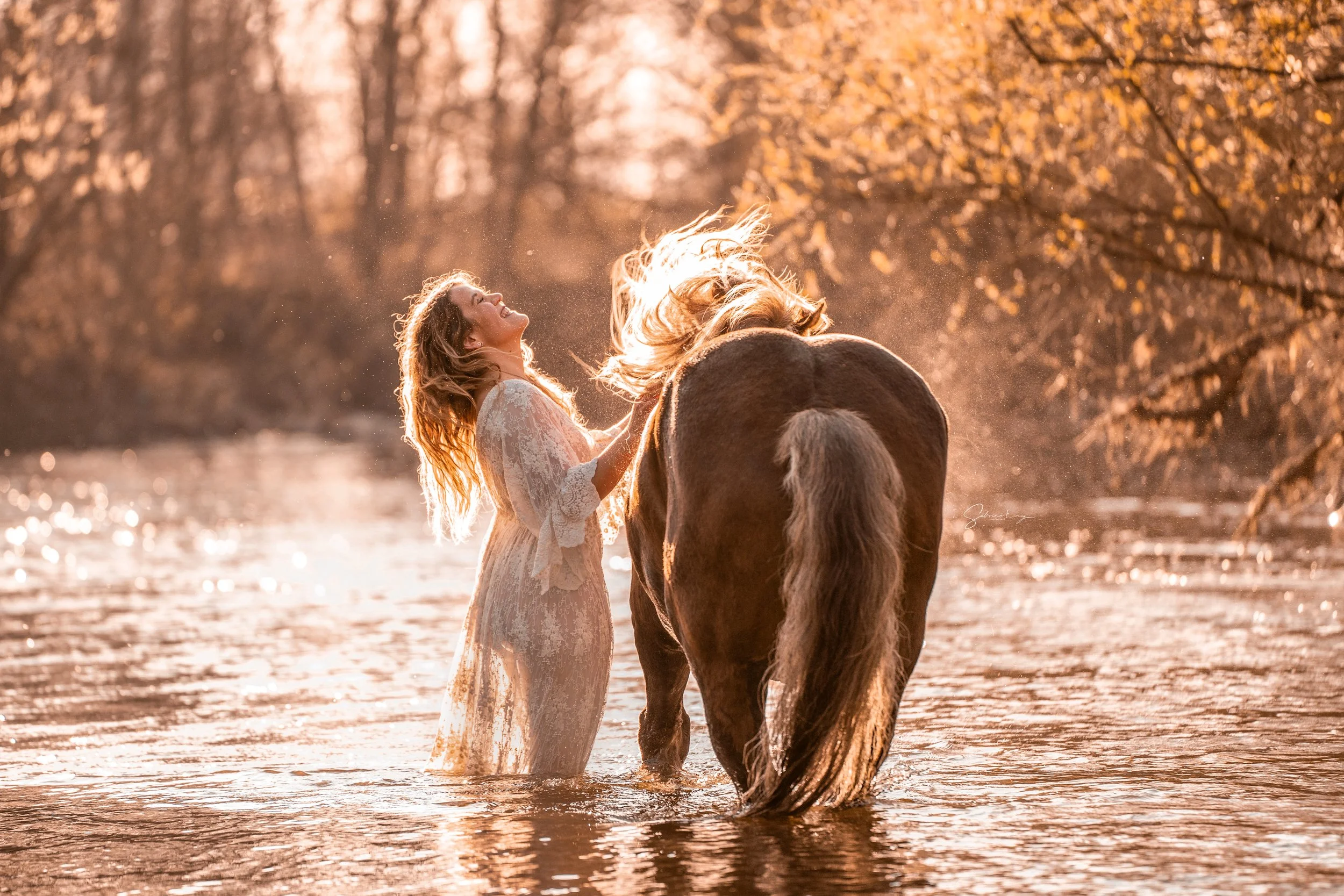 Eine Frau in einem weißen Spitzenkleid steht lächelnd mit wehenden Haaren im seichten Wasser neben einem Pferd bei Sonnenuntergang in einem Waldgebiet mit Bäumen und goldenem Herbstlaub.
