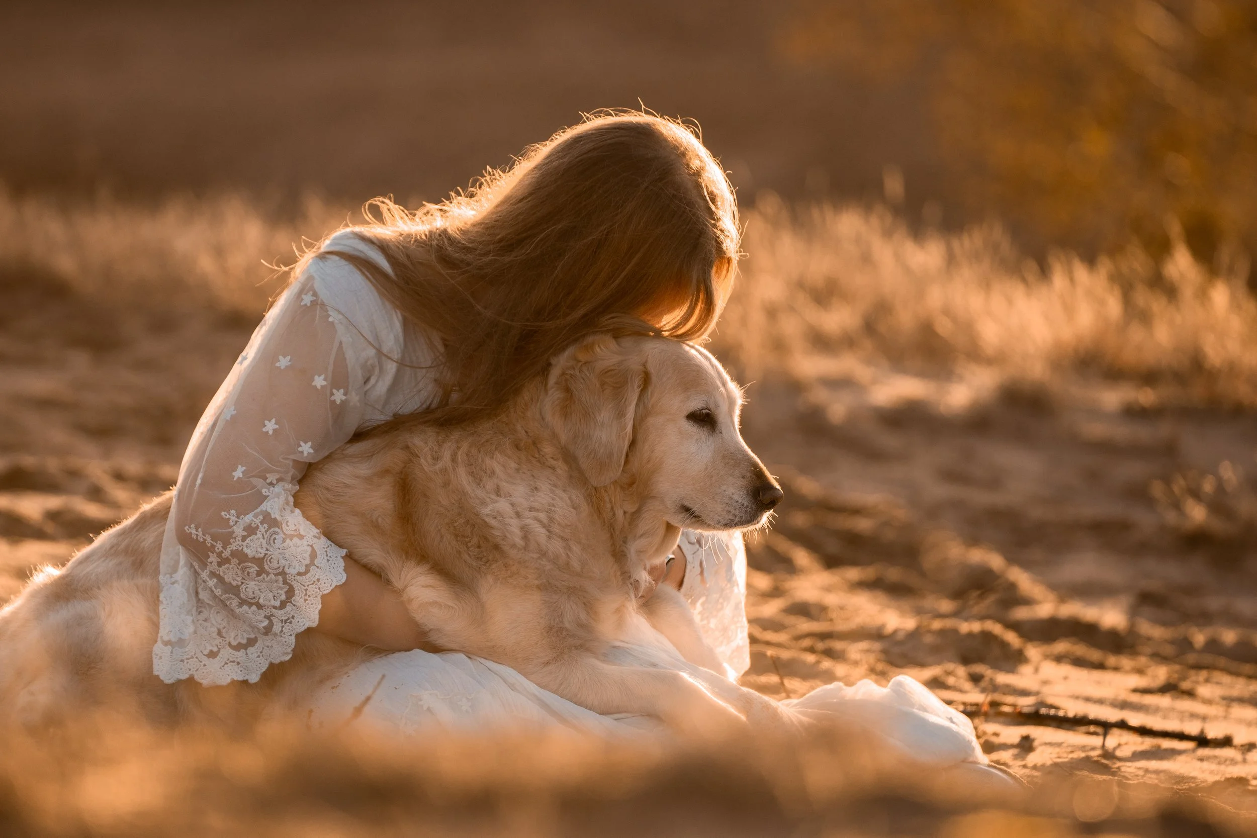 A woman in a white dress with lace details hugging a Golden Retriever puppy outdoors during sunset.