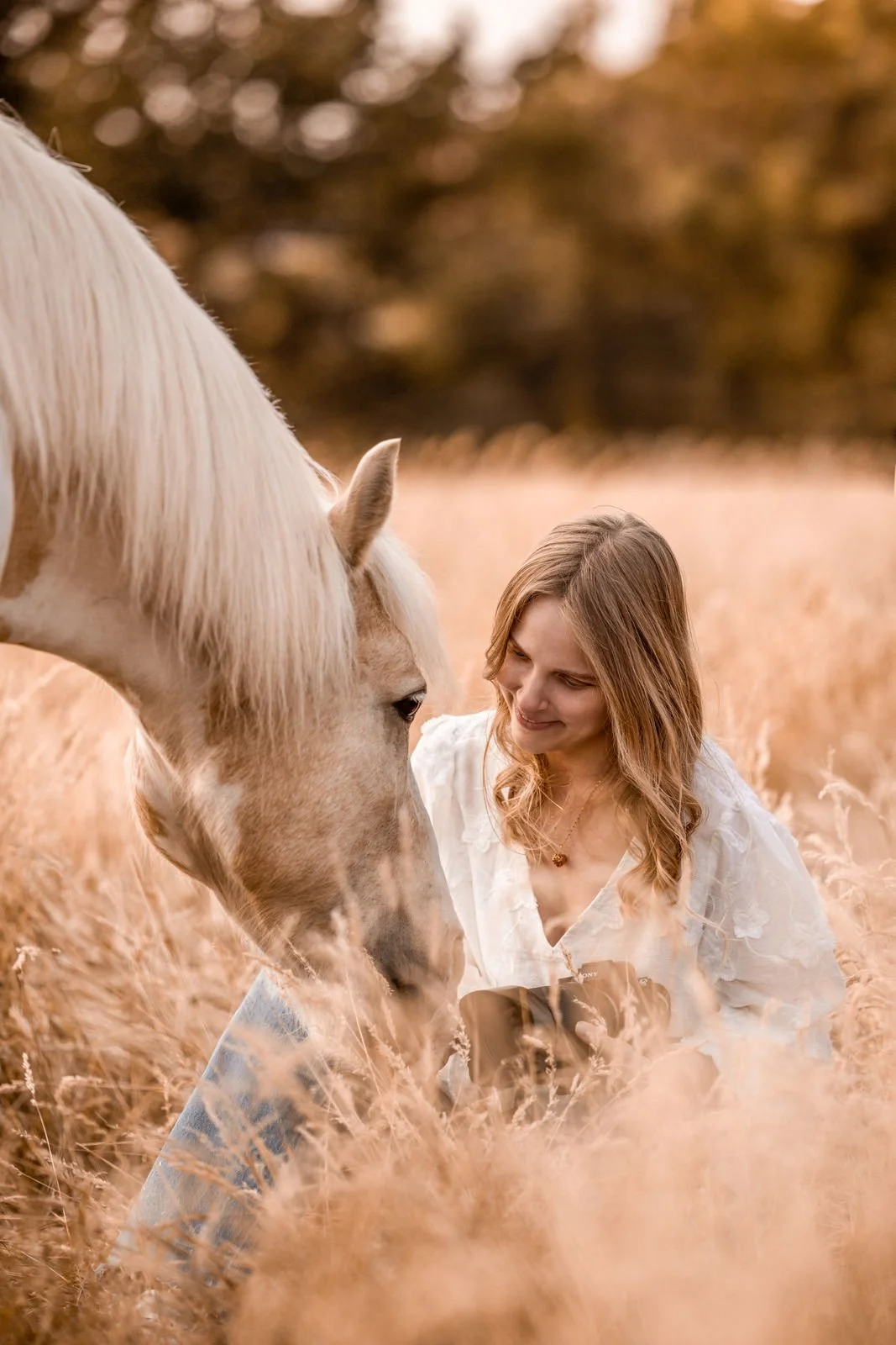 Eine Frau mit langen Haaren und einem weißen Kleid sitzt in einem goldenen Feld neben einem hellen Pferd und schaut es liebevoll an.
