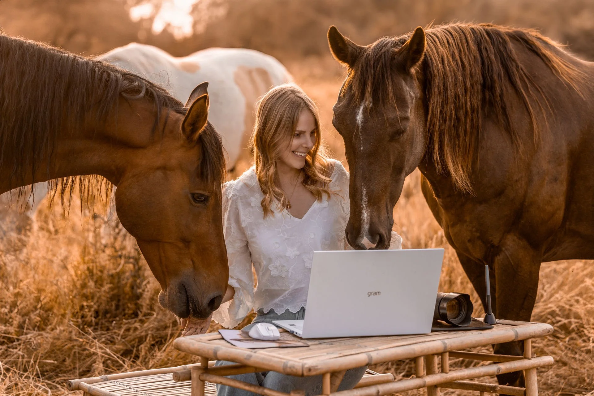 Eine junge Frau mit blonden, lockigen Haaren arbeitet im Freien auf einem Holztisch an einem Laptop, umgeben von drei Pferden im Sonnenuntergang.