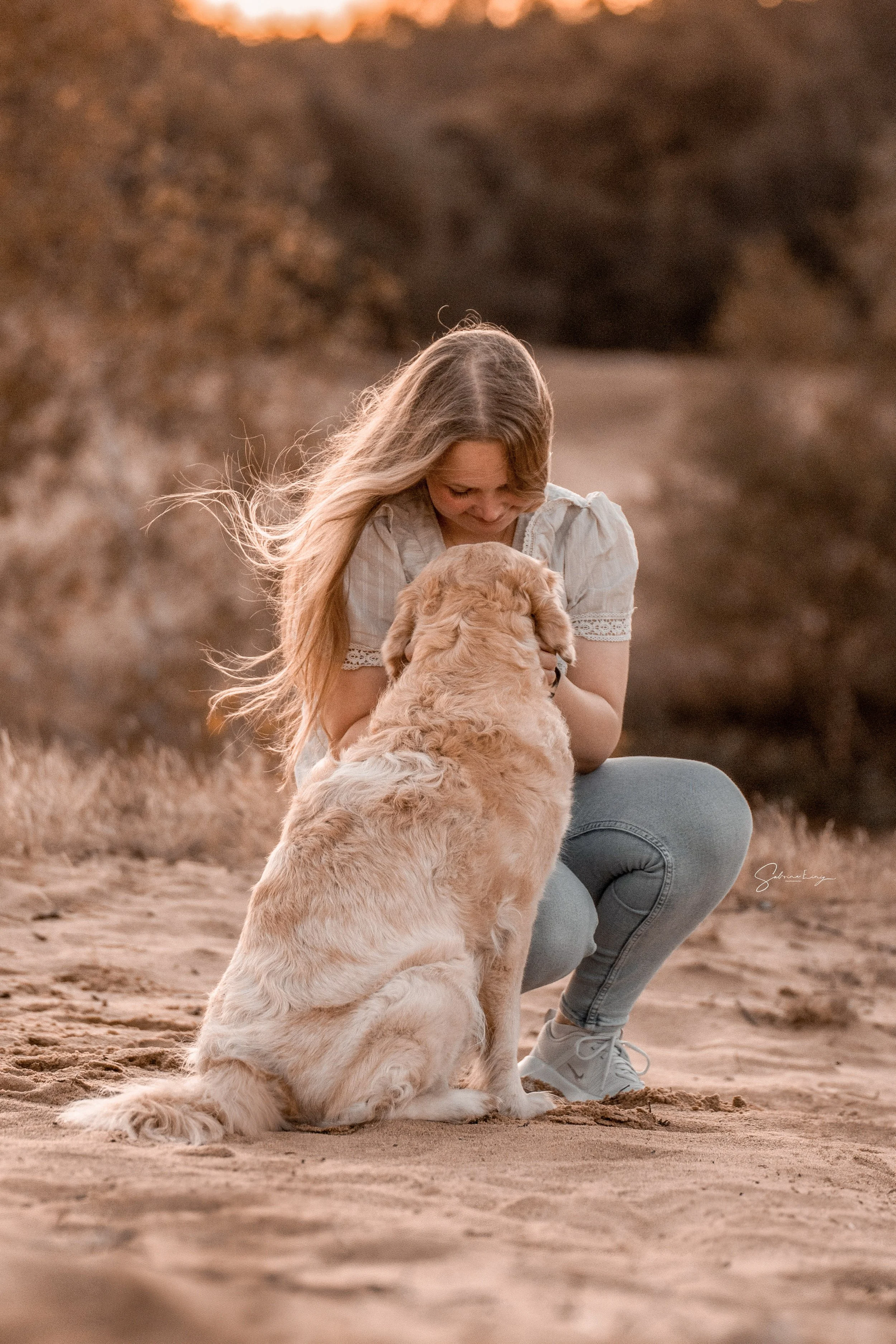A young woman with long blonde hair kneeling on a sandy beach, holding a golden retriever puppy and looking down at it with affection during sunset.