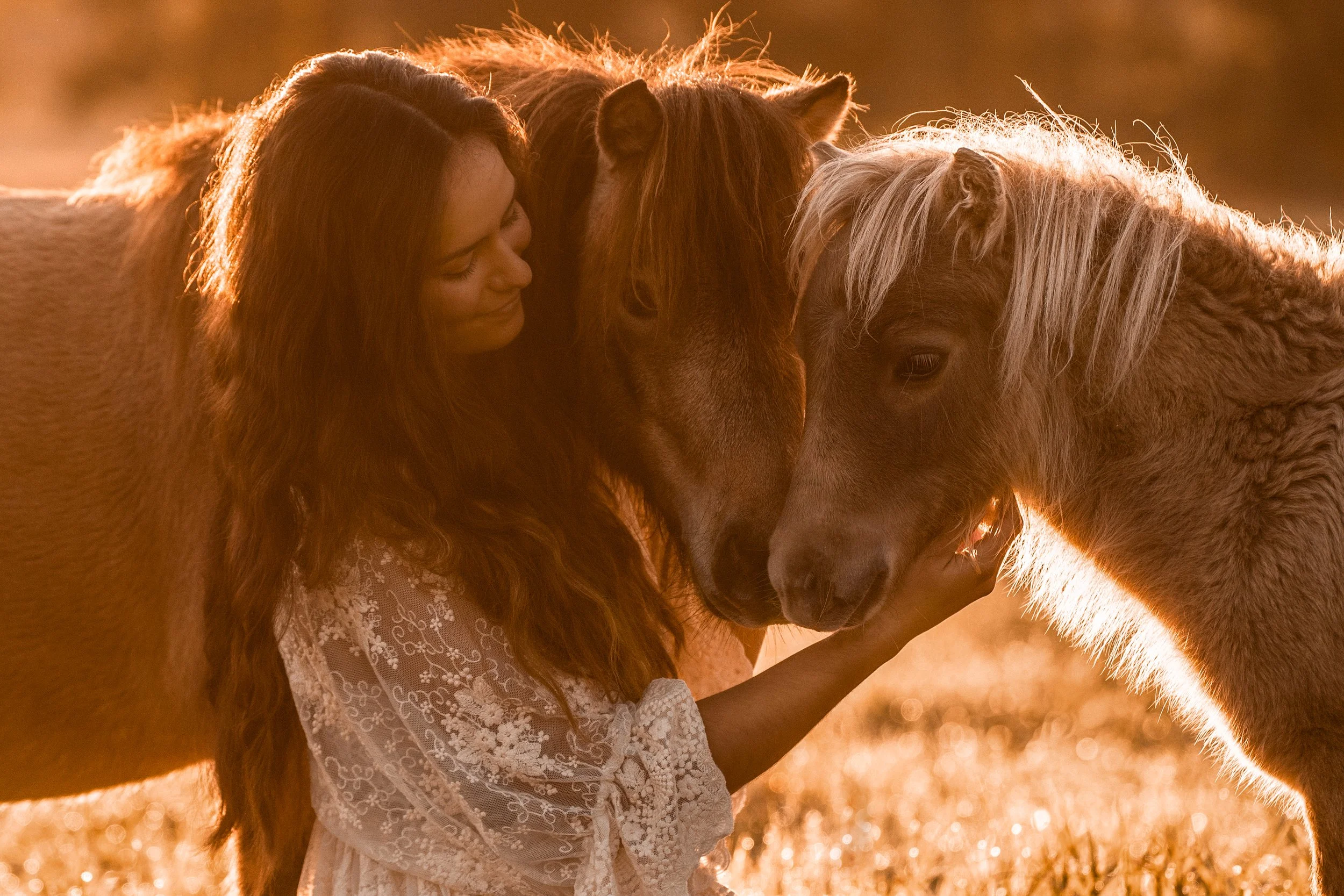 Eine Frau mit dunklem Haar in einem weißen, durchbrochenen Kleid streichelt bei Sonnenuntergang im Freien zwei kleine Ponys mit erdfarbenem Fell.