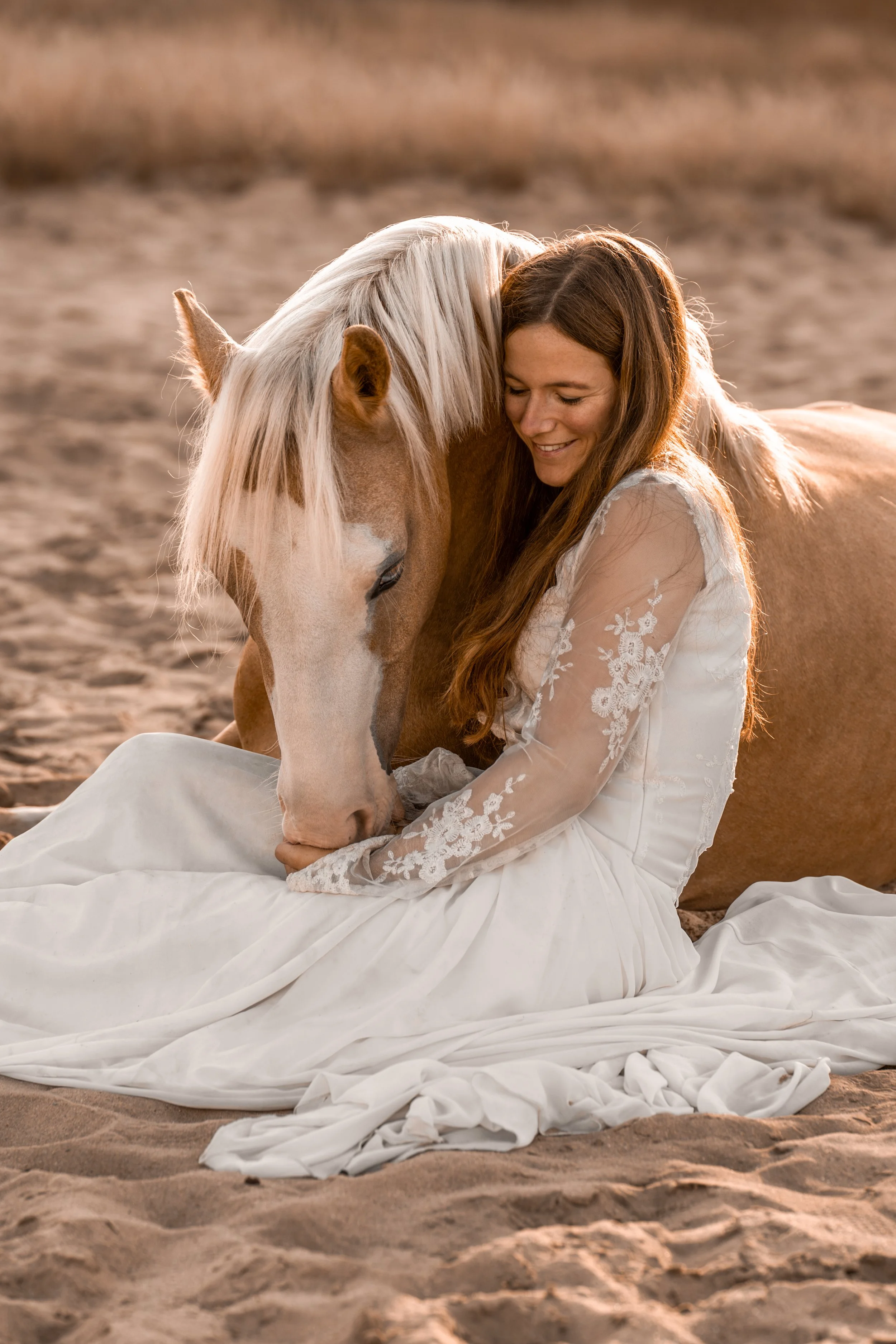 Eine Frau in einem weißen Spitzenkleid sitzt am Strand im Sand und umarmt ein Palomino-Pferd mit weißer Mähne; beide haben ihre Köpfe aneinander gelehnt und lächeln sich während der goldenen Stunde warm an.