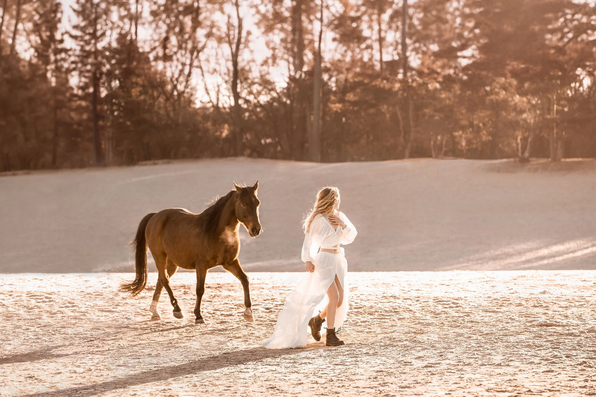 Eine Frau in einem weißen Kleid und schwarzen Stiefeln geht in der Nähe eines Pferdes über ein sandiges Feld, im Hintergrund sind Bäume zu sehen, während die Sonne untergeht.