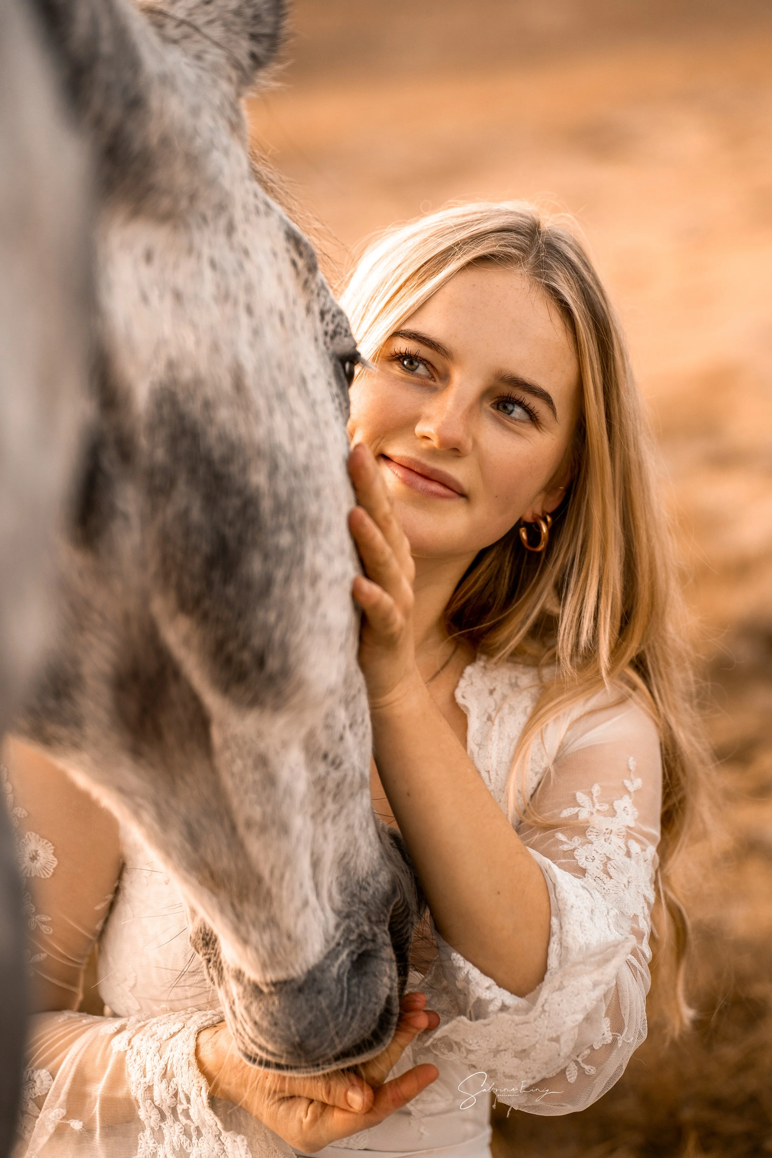 Eine junge Frau mit langen blonden Haaren und Creolen, die sanft das Gesicht eines Pferdes streichelt und es liebevoll ansieht, in einer natürlichen Außenkulisse mit warmer Beleuchtung.