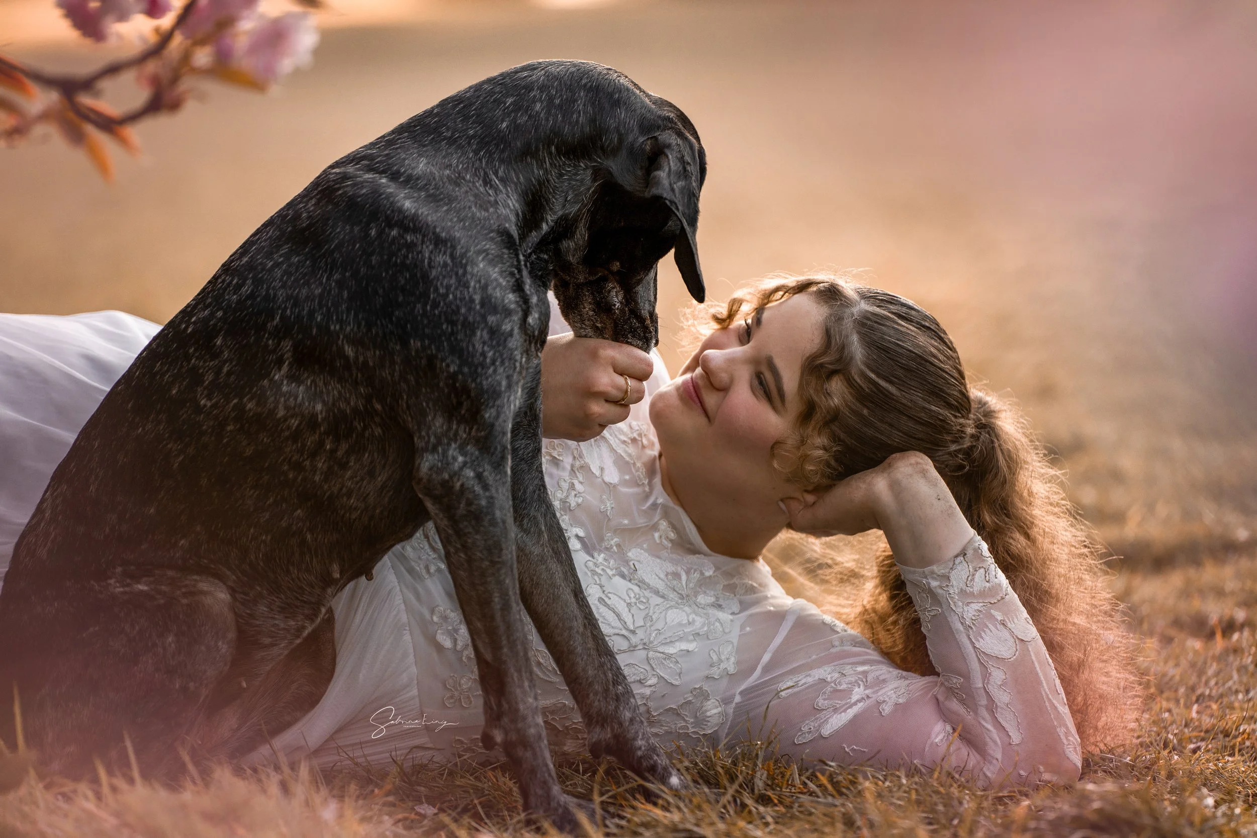 A woman with curly hair and a white lace dress lying on the grass, smiling as a large black and grey dog leans over her, touching her face with its paw.